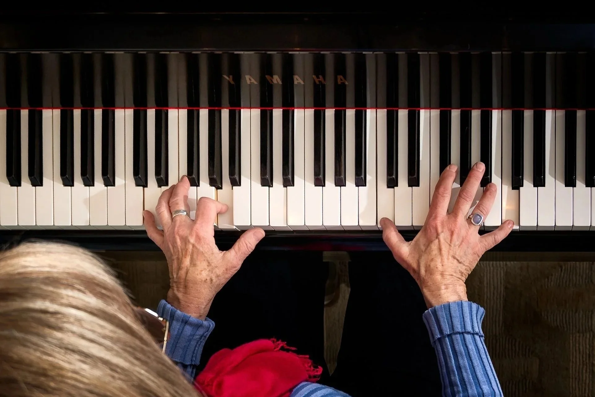 Overhead view of an elderly person playing a Yamaha piano, with hands on the keys, wearing a blue sweater and rings. Another person's head with blonde hair is visible in the foreground.