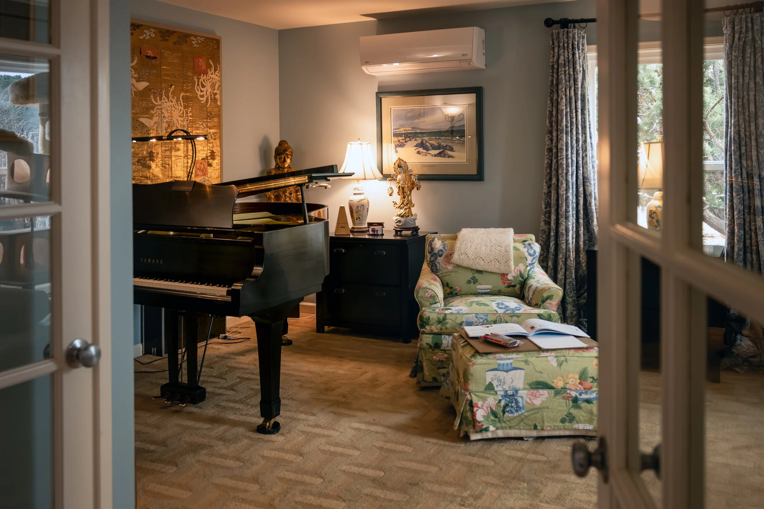 A cozy living room with a black grand piano, a floral upholstered armchair with a pillow, a dark wooden side table, and decorative items including a statue, a lamp, and framed artwork. Large windows with patterned curtains allow natural light to brighten the space.