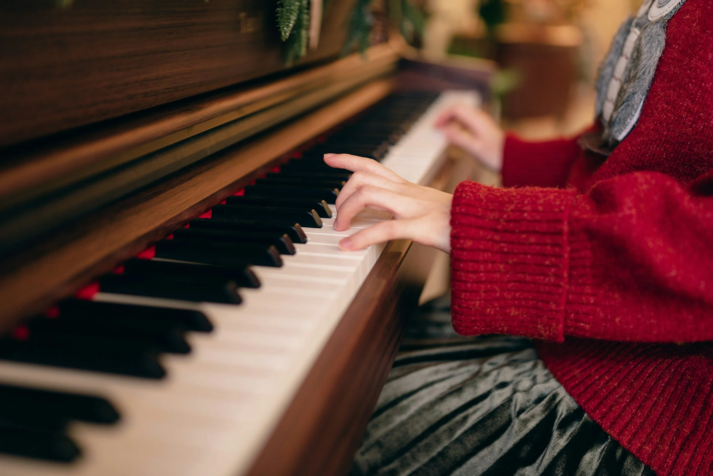 Close-up of a person playing a wooden upright piano with black and white keys. The person is wearing a red knitted sweater and patterned pants.