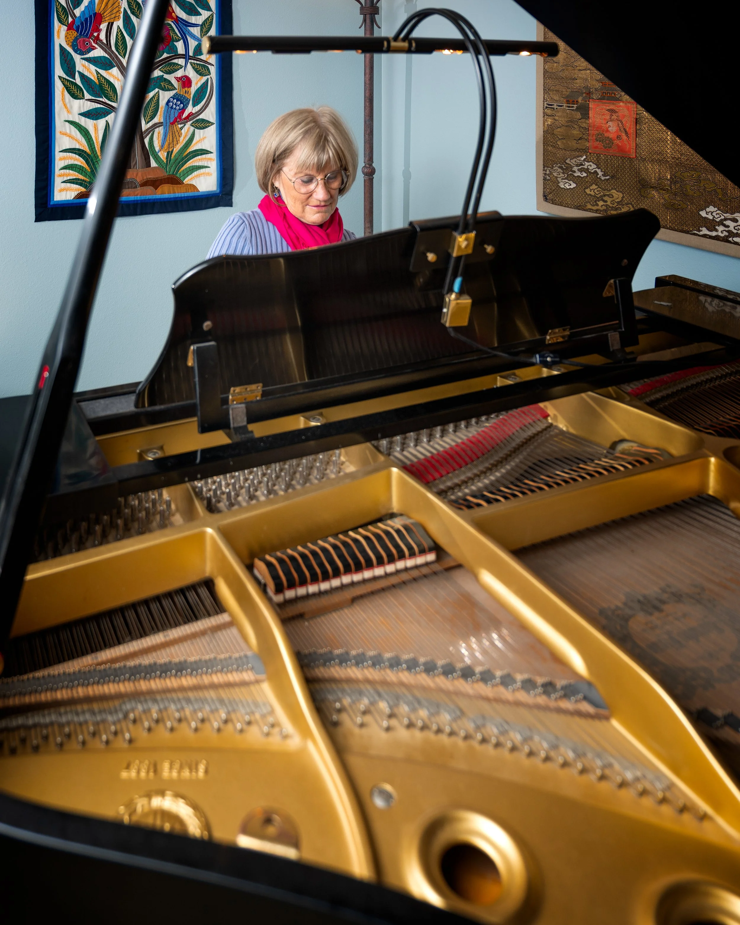 A woman with gray hair wearing glasses, a pink scarf, and a striped shirt playing a grand piano indoors, with colorful tapestries hanging on the walls.