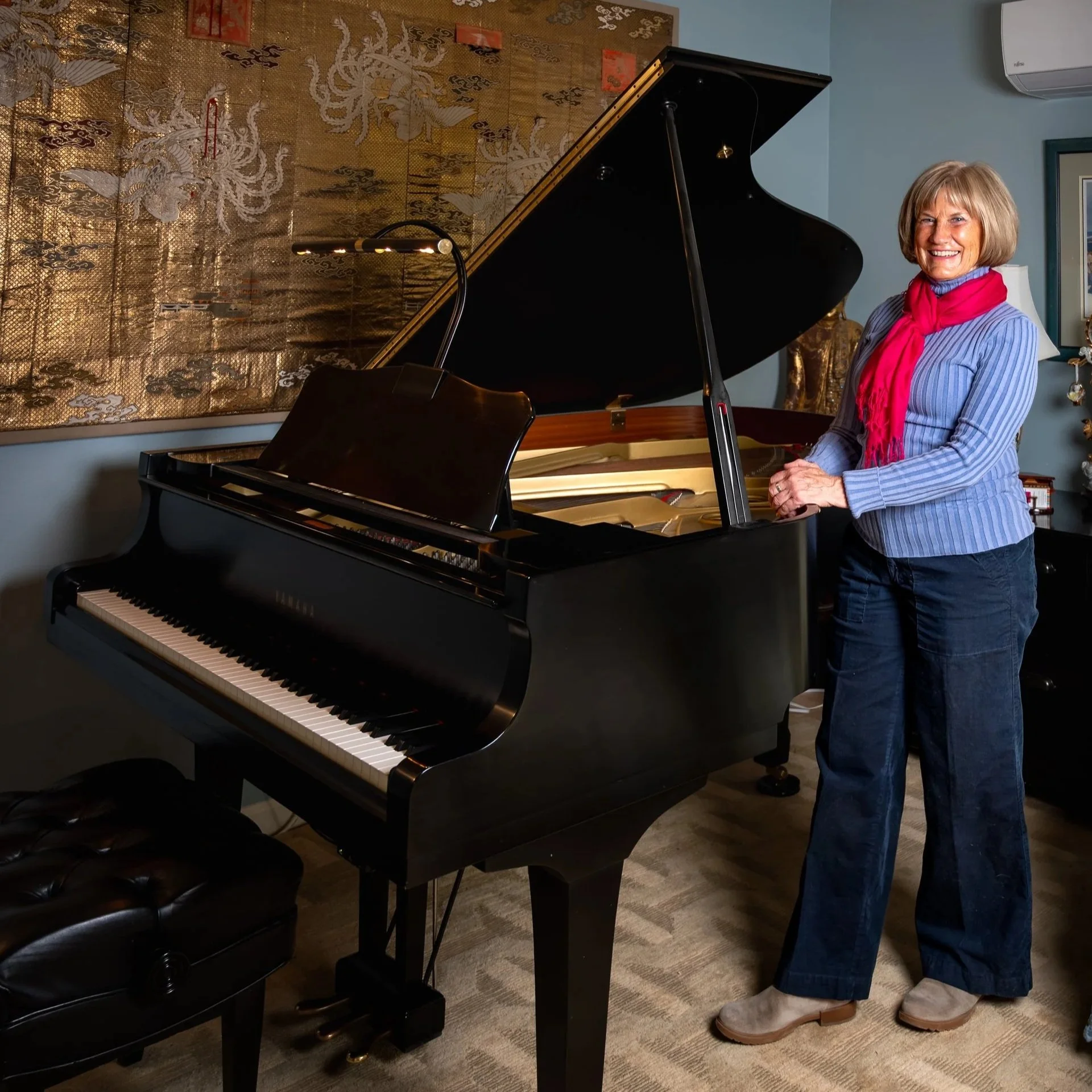 Woman standing next to a grand piano in a room with a decorative wall hanging of a dragon and a lamp in the background. The woman is smiling, wearing a striped shirt with a red scarf, jeans, and beige shoes.