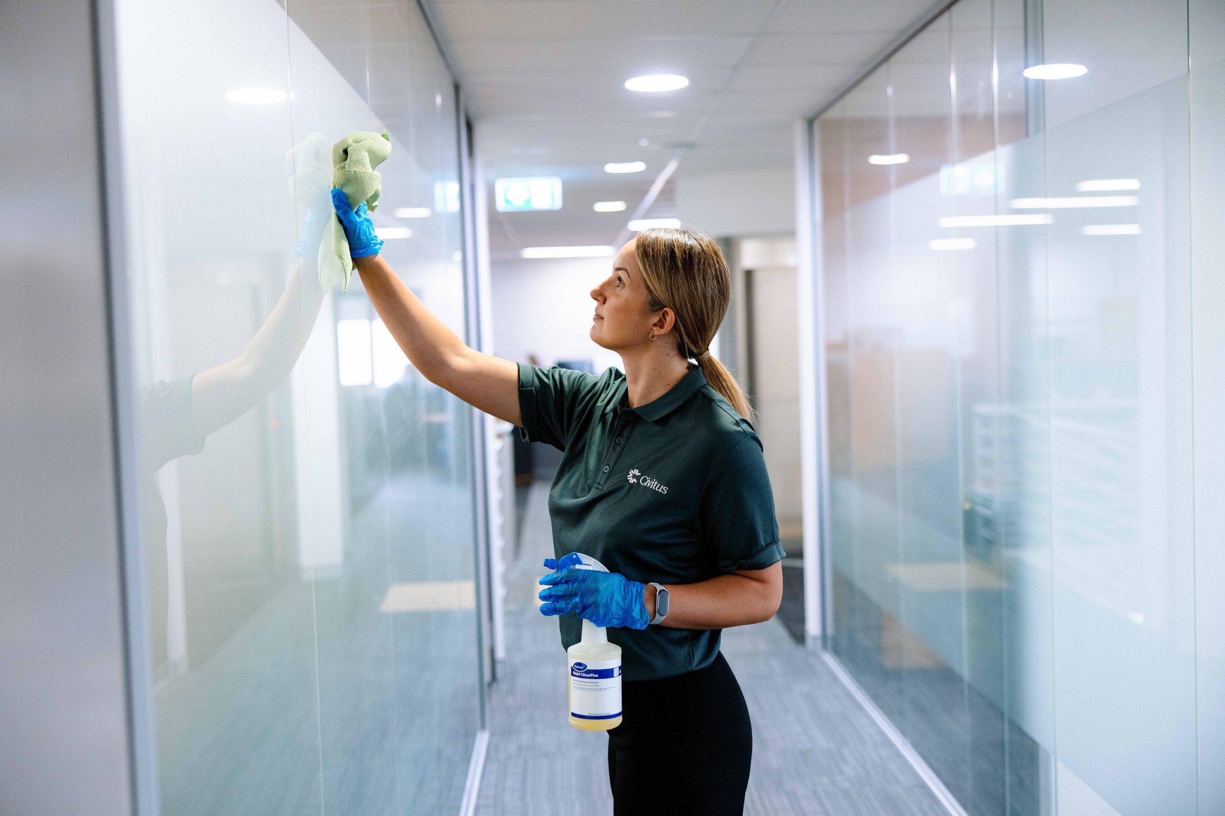 A woman cleaning a glass wall in a modern building hallway. She is wearing a dark shirt, blue gloves, and a smartwatch, and is holding a spray bottle. The hallway has reflective glass walls and ceiling lights.