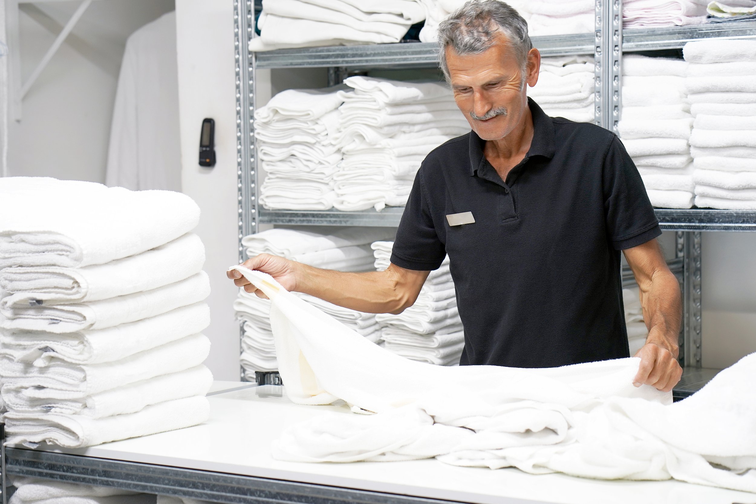 A man with gray hair and a mustache, wearing a black polo shirt, is folding a white towel in a laundry room with shelves filled with neatly folded white towels and linens.