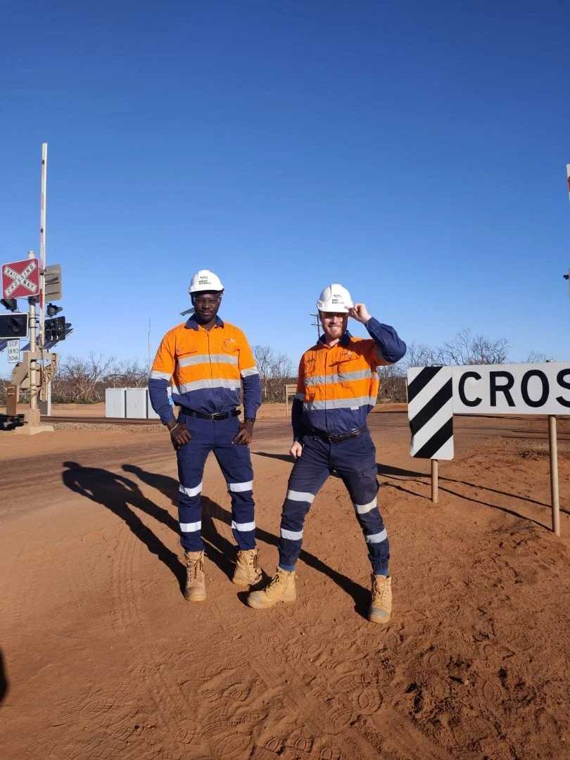 Two construction workers in safety gear, standing on a dirt road near a railroad crossing, wearing orange and navy uniforms and hard hats, with clear blue sky in the background.