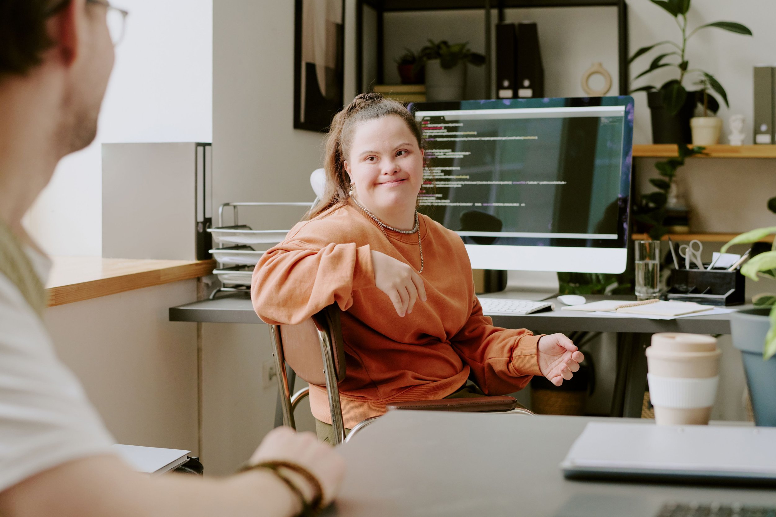 A woman with long hair and pearl earrings smiling at a man in an office setting, with a computer monitor displaying code behind her.