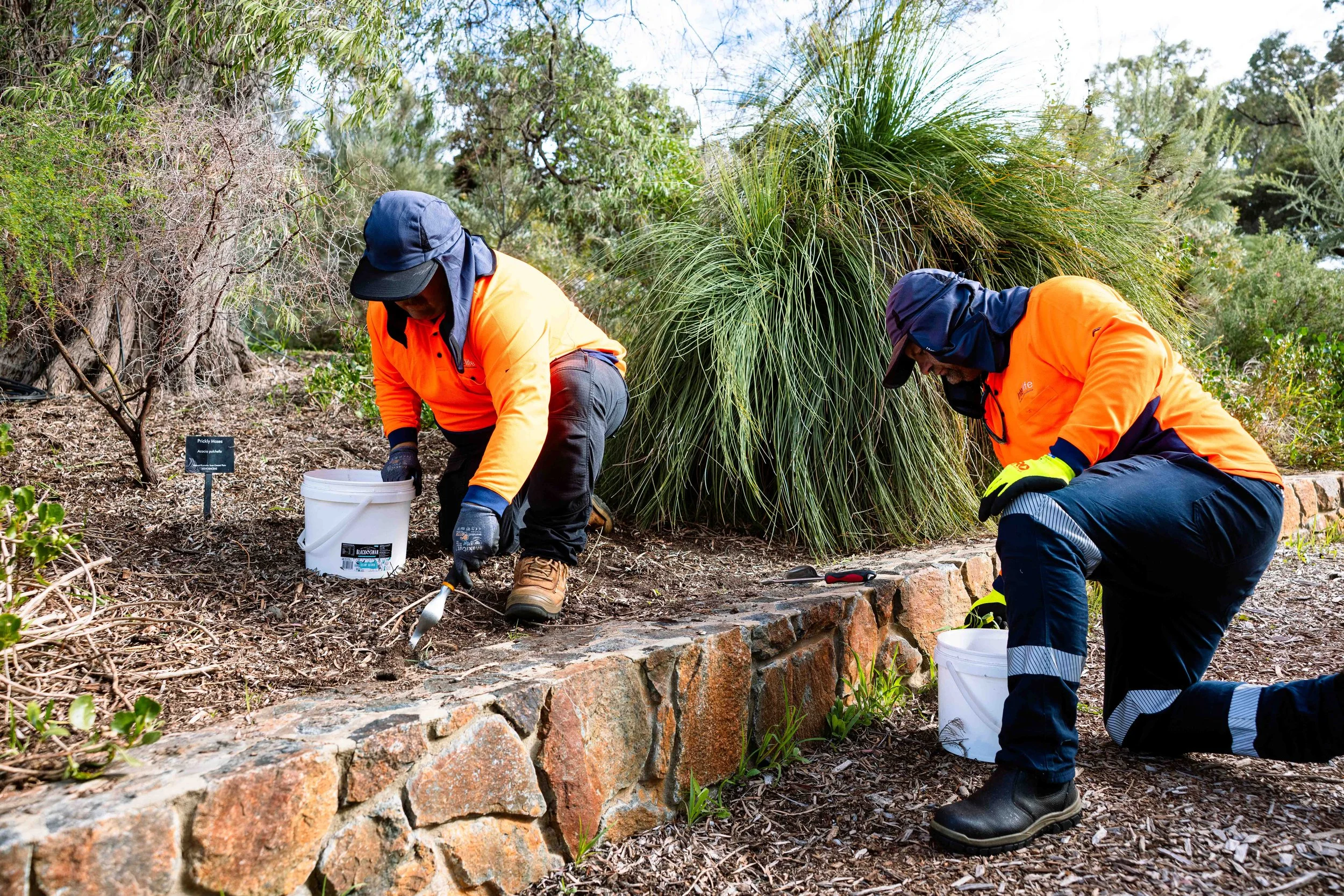 Two workers installing or maintaining a stone border or wall in a garden, with greenery and trees in the background.