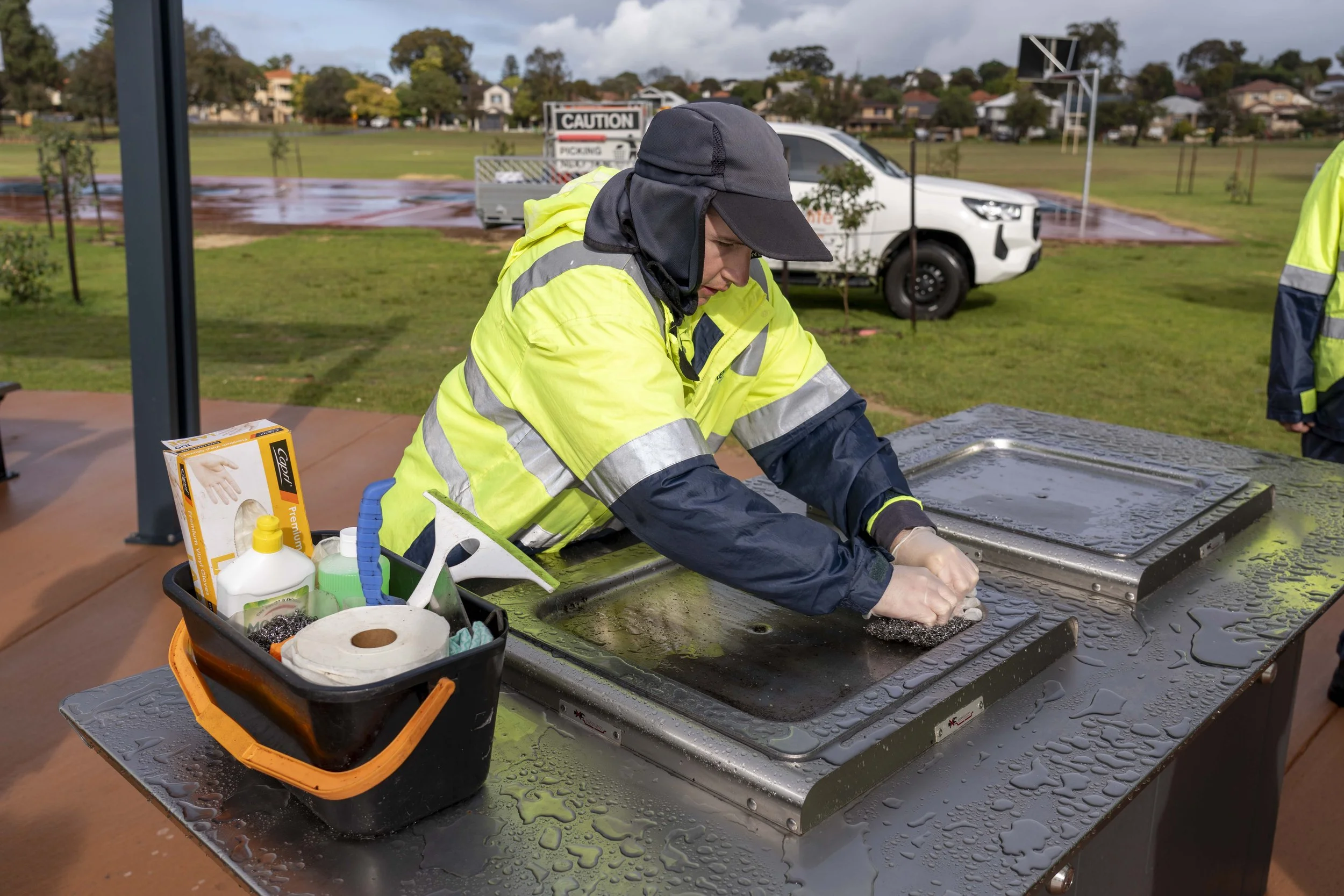 A person wearing a high-visibility yellow jacket and a dark hat is cleaning a black surface on a table outdoors. There are cleaning supplies and paper towels on a tray nearby. In the background, there is a sports court, a white vehicle, and a neighborhood of houses with trees under a cloudy sky.