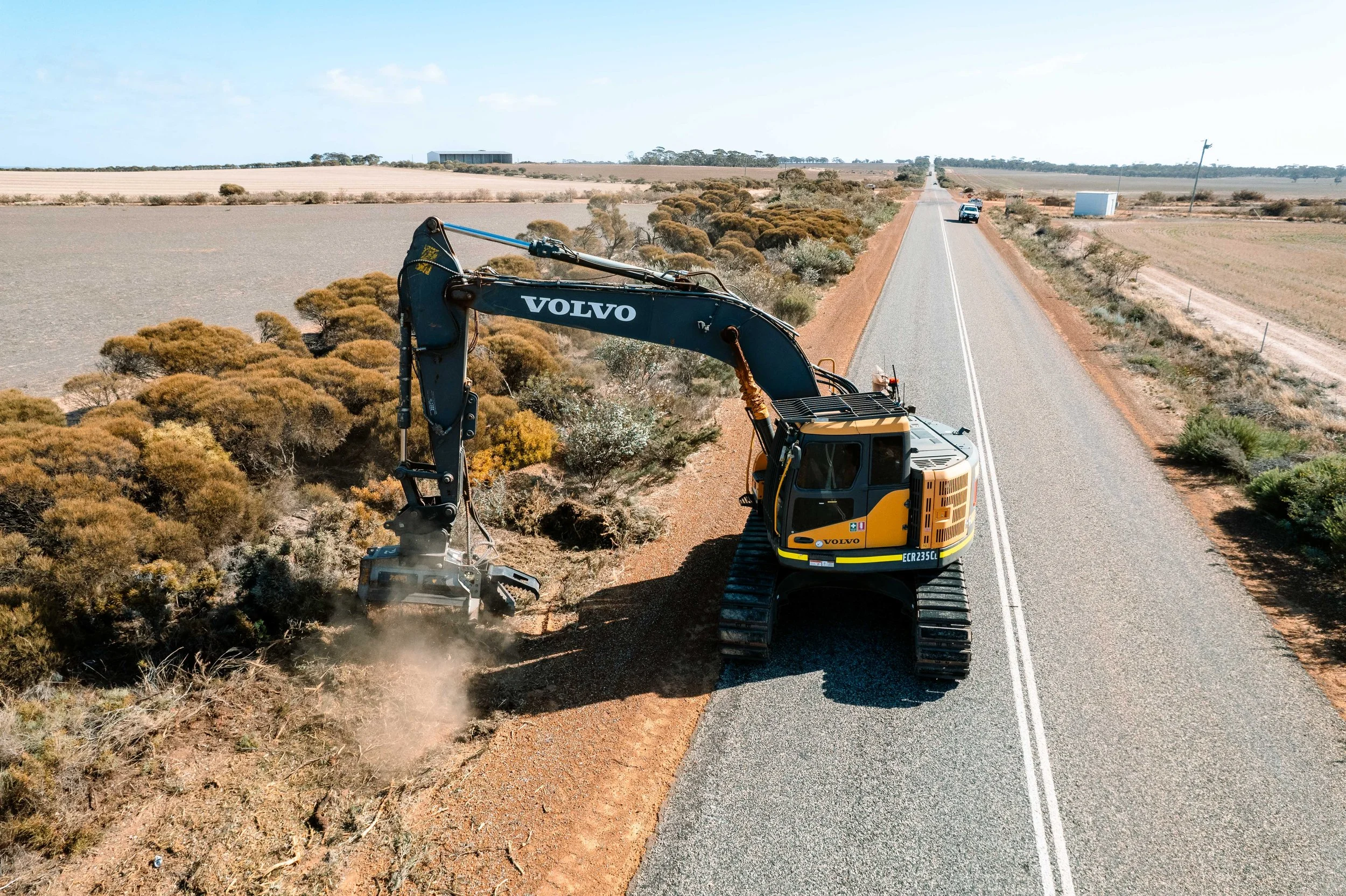 A Volvo construction excavator working on the side of a rural road, clearing or excavating dirt and vegetation in a dry, open landscape with fields and minimal vegetation.