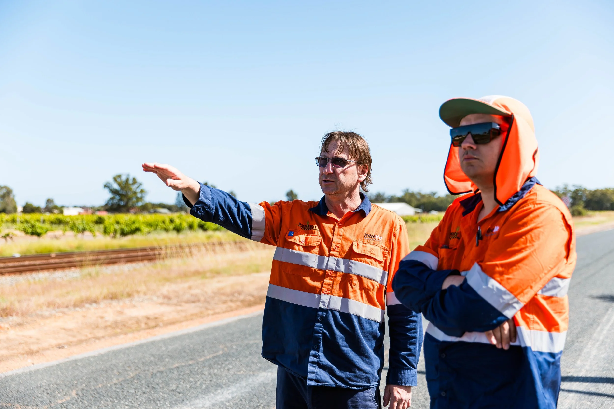 Two men in orange uniforms, one with glasses and one wearing a hat and sunglasses, discussing possibly near railway tracks outdoors on a sunny day.