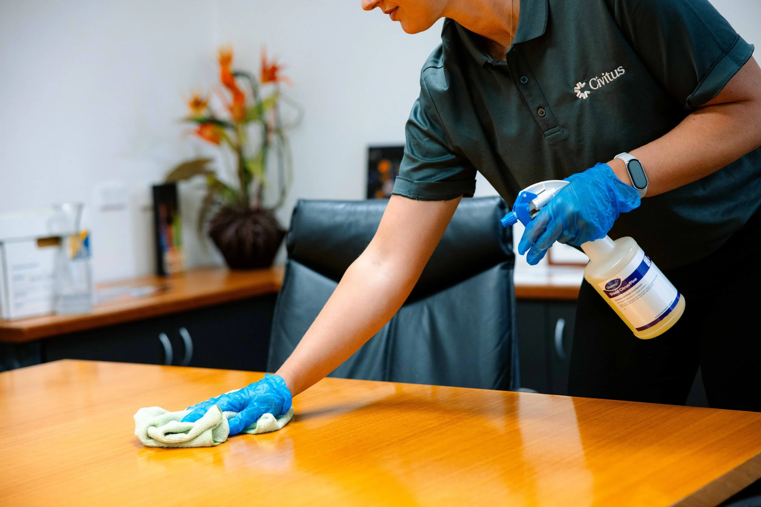 A person wearing a dark polo shirt with the logo 'Civitus' and blue gloves is disinfecting a wooden table with a spray bottle, with a tissue or cloth on the table in an office setting.