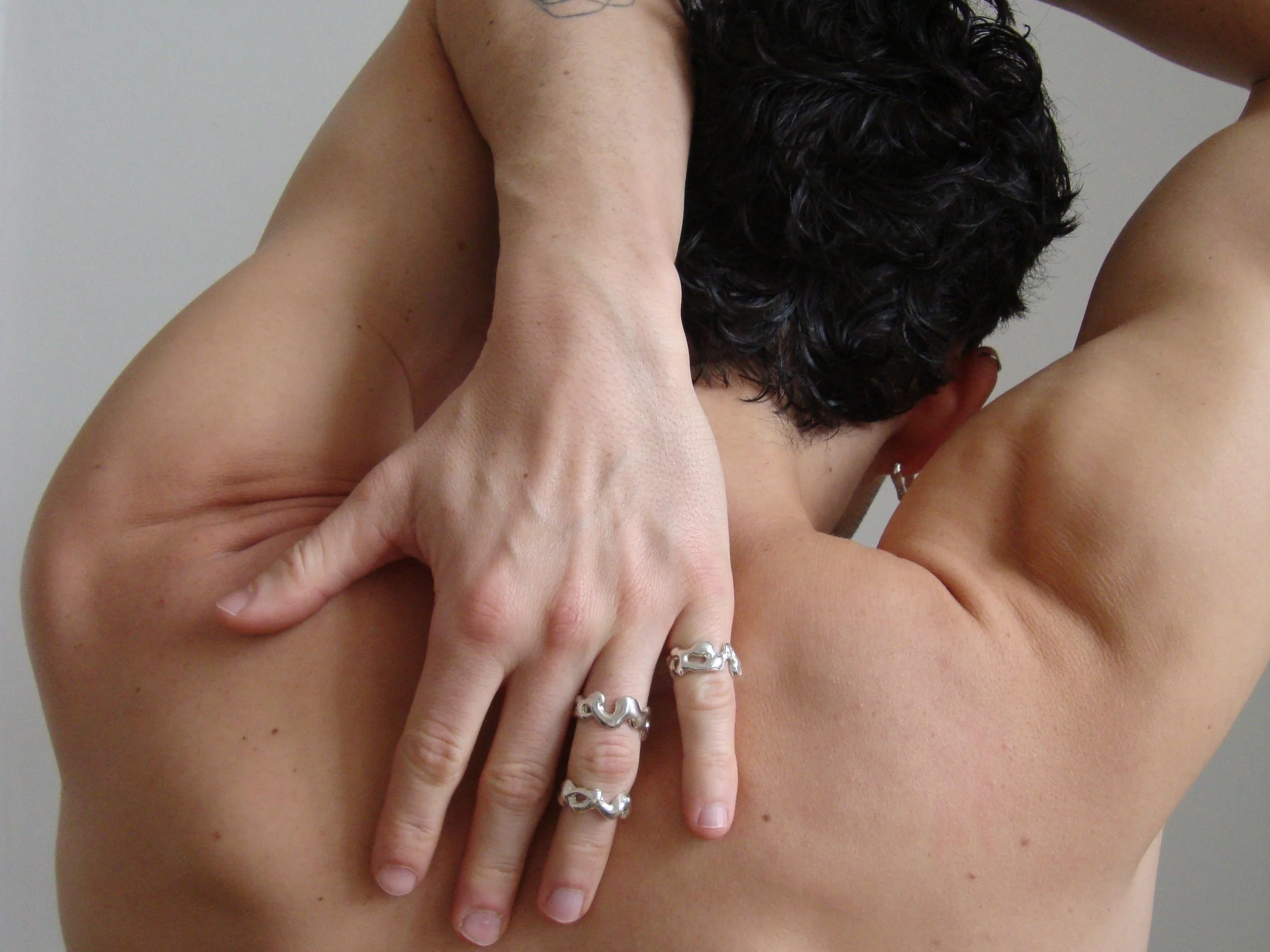 A person with short dark curly hair is holding their arm behind their head and wearing multiple rings.