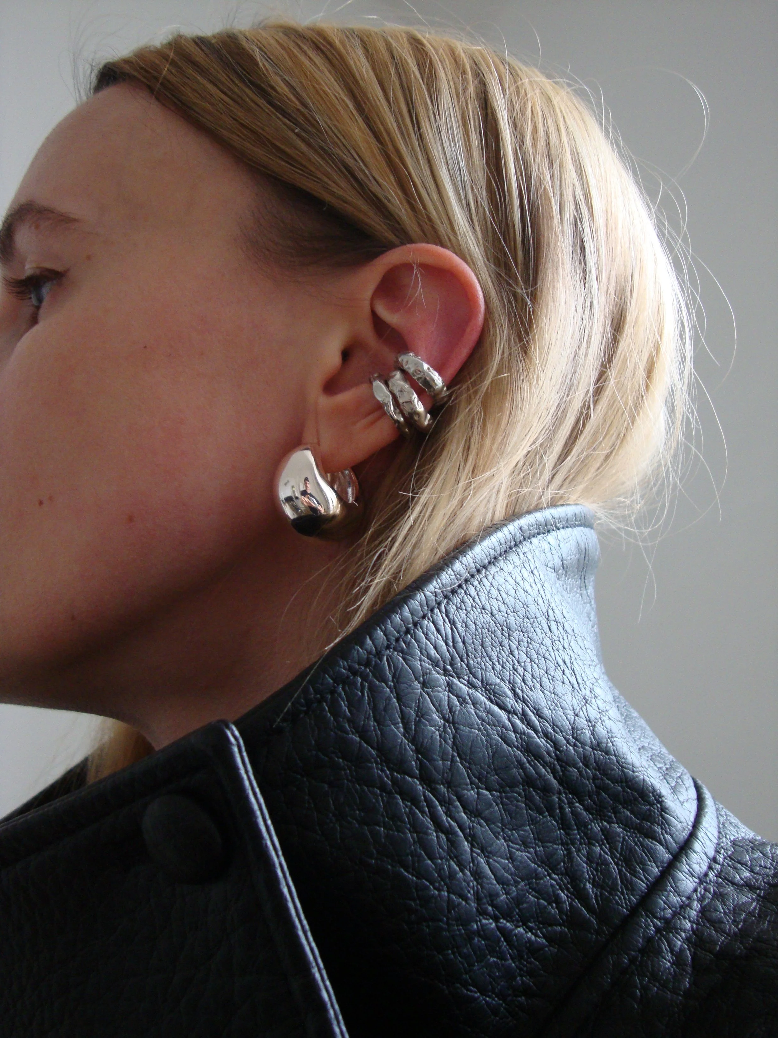 Close-up side view of a woman's head and shoulder, showing her blonde hair, multiple silver earrings, and black leather jacket.