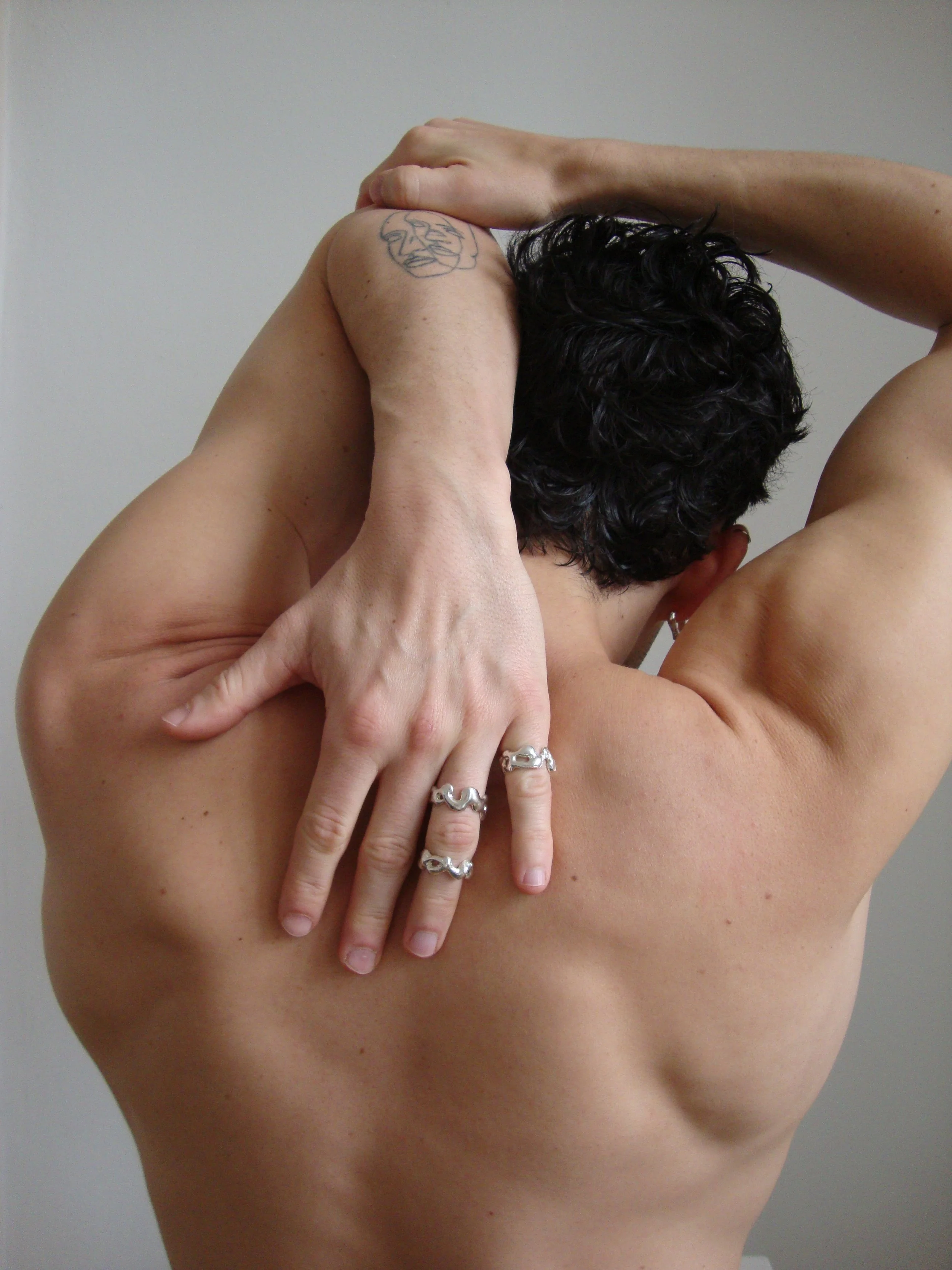 A person with short dark curly hair is holding their arm behind their head and wearing multiple rings.