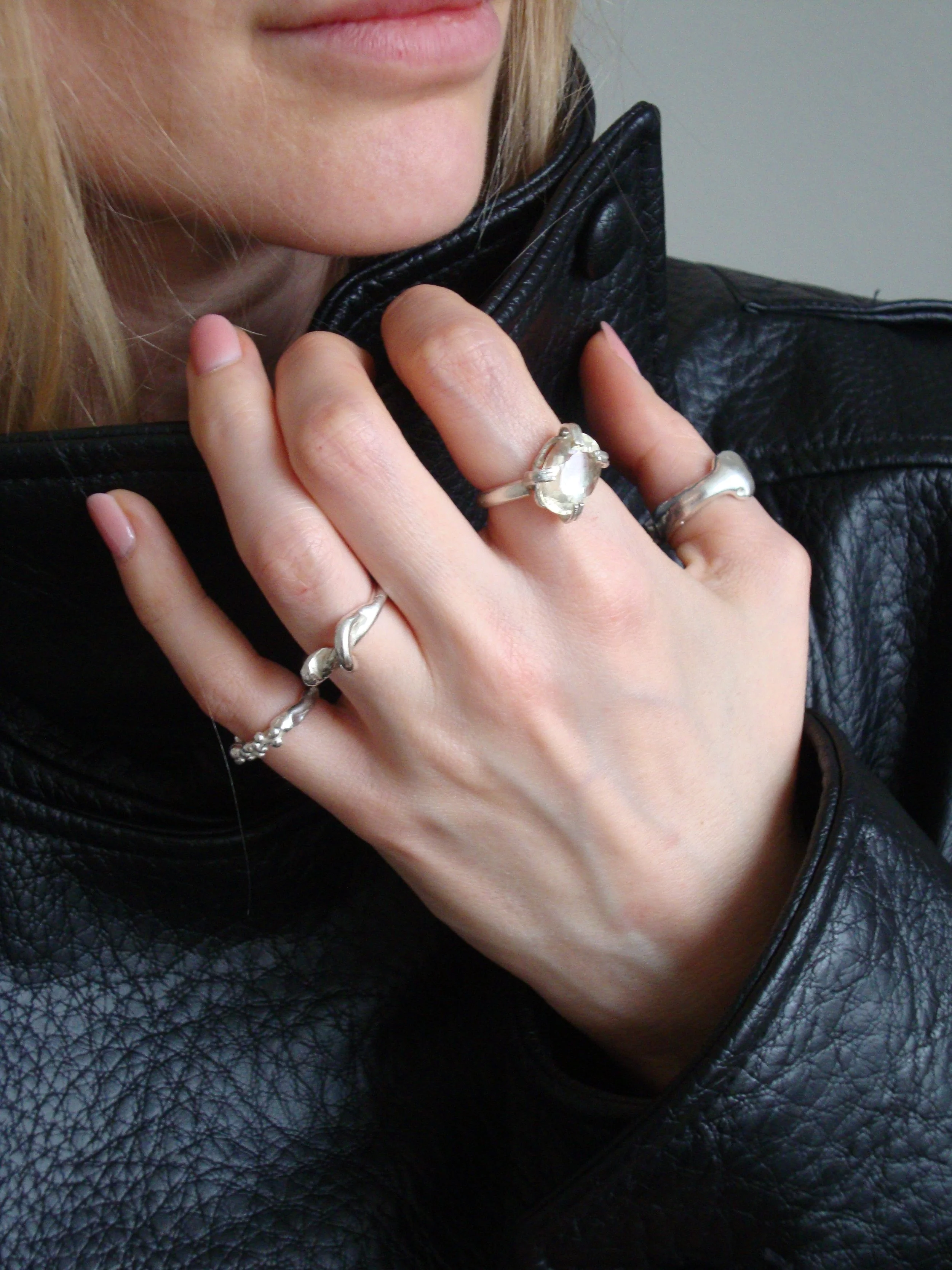 A woman wearing a black leather jacket with her hand near her neck, showing multiple silver rings on her fingers.