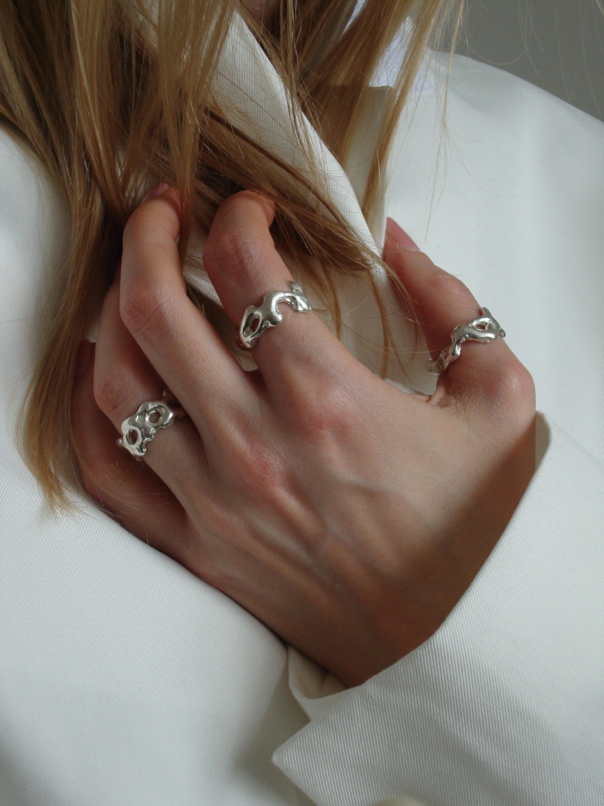 Close-up of a person's left hand with three silver skull rings, holding what appears to be a strand of light brown hair, against a white fabric background.