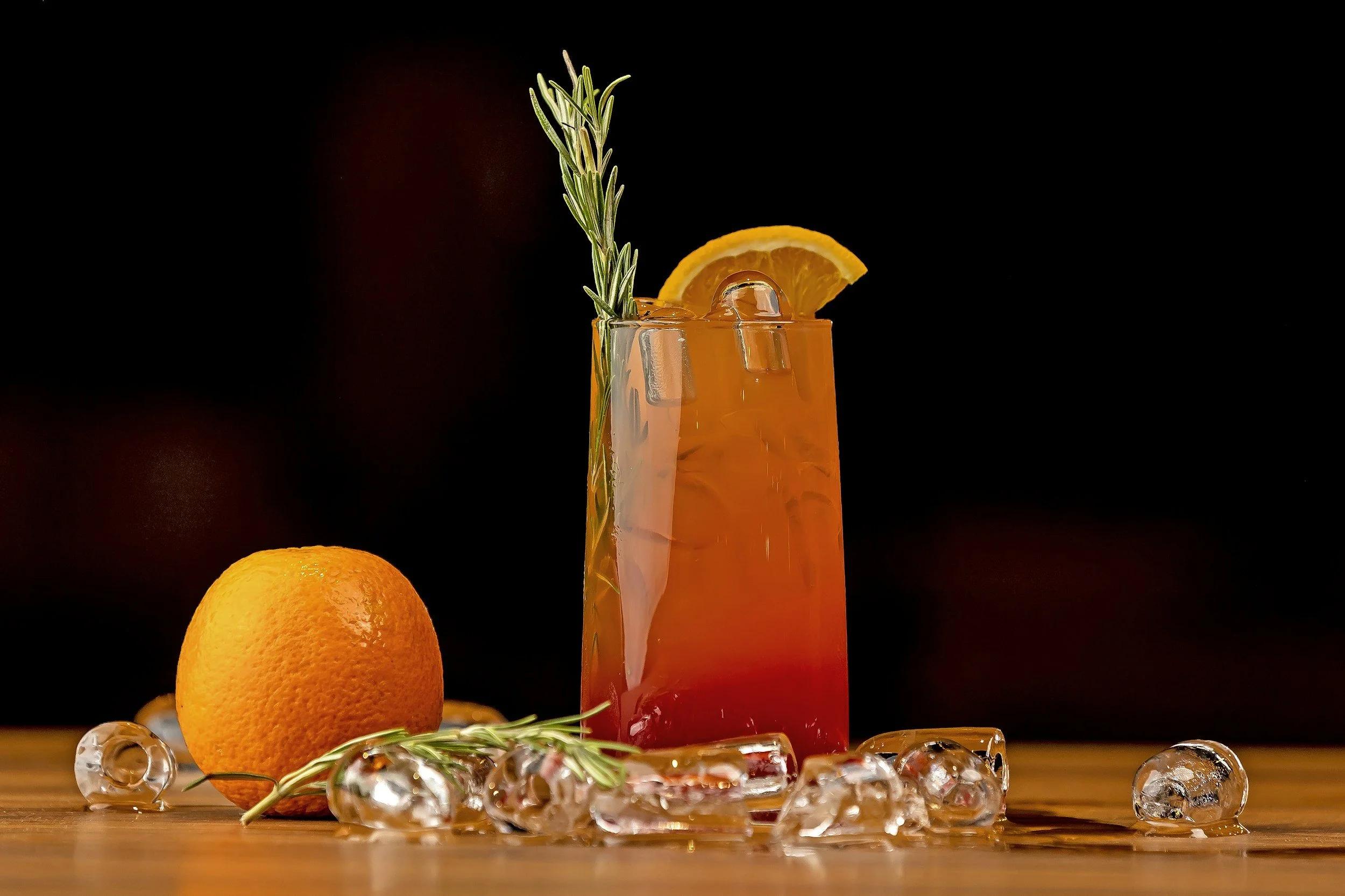 A tall glass of orange-colored beverage garnished with an orange slice and sprig of rosemary, with ice cubes inside, on a wooden surface with scattered ice cubes and an orange fruit nearby, against a black background.