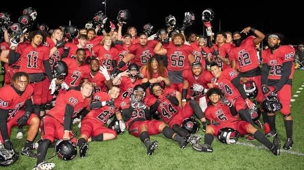 A group of football players in red uniforms and helmets posing on a field at night. Some players are sitting, others standing, with helmets raised in celebration.