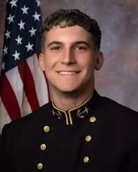 Young man in military uniform smiling, American flag in background.