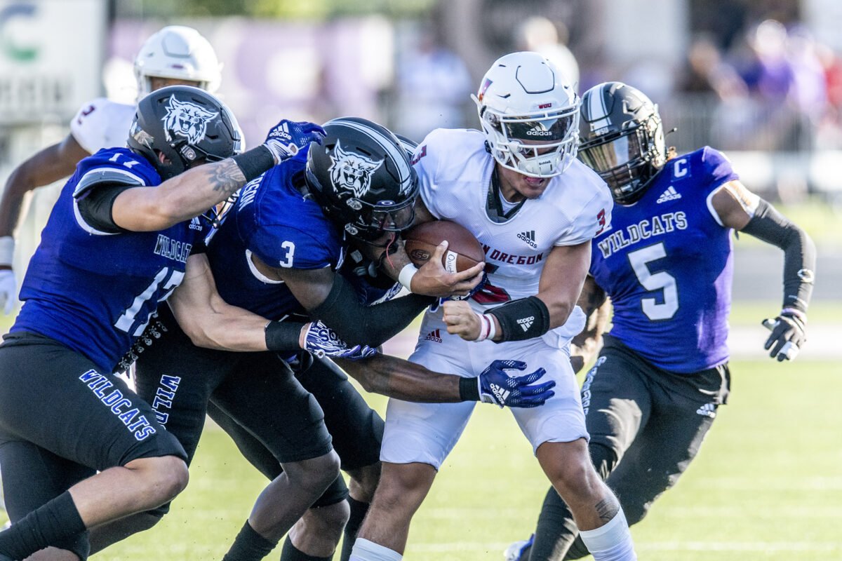 A football game with a player from the Oregon Ducks in white uniform carrying the ball while being tackled by three players from the Wildcats team in blue uniforms.