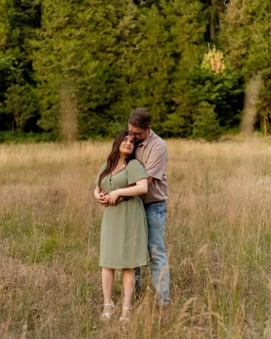 A couple standing in a grassy field, with the man holding the woman from behind, surrounded by trees.