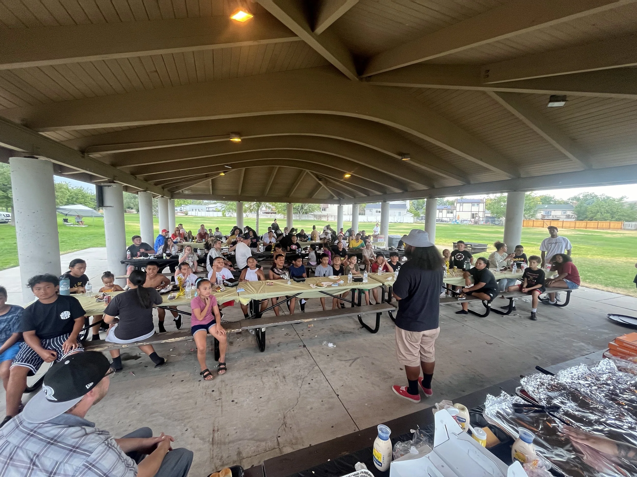 A large group of children and adults sitting under a pavilion at a gathering, with food on tables, on a sunny day in a park.