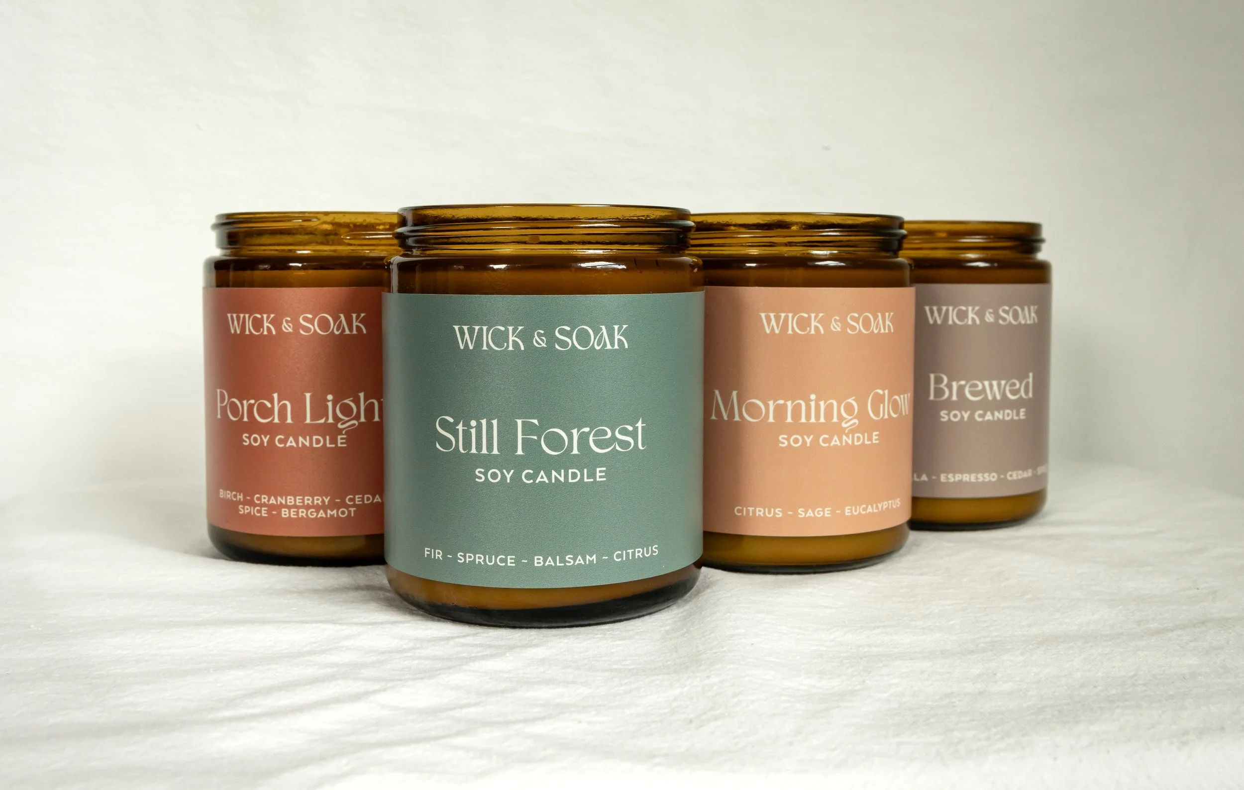 A row of five amber glass candles labeled Wick & Soak soy candles. The candles have pastel-colored labels with different scents, including Porch Light, Still Forest, Morning Glow, and Brewed.