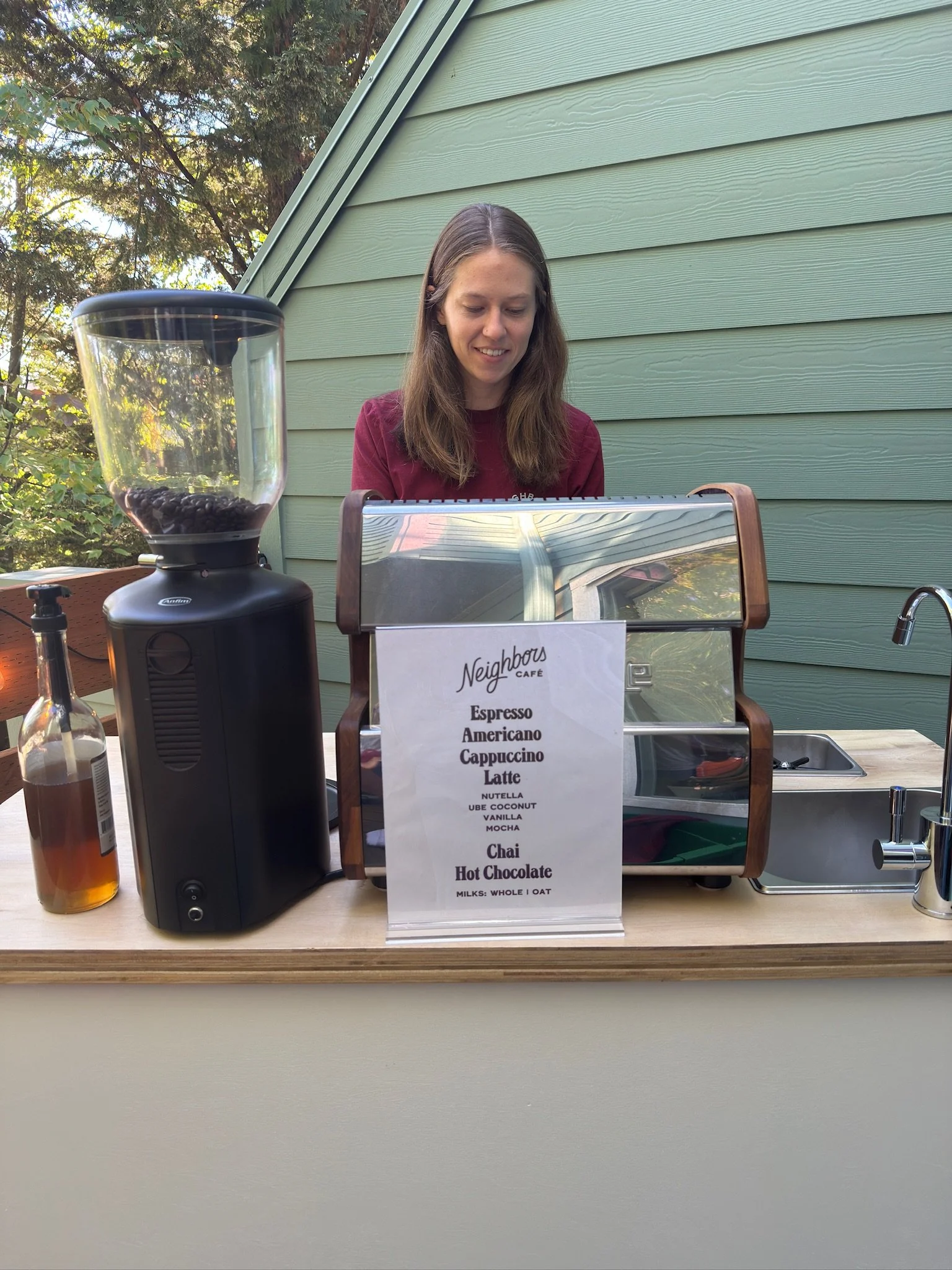 Outdoor cafe stand with a woman smiling behind a counter. There is a menu on the front listing various drinks like espresso, americano, cappuccino, latte, and hot chocolate, with milk options. A large coffee grinder is on the left, and a bottle of flavor syrup is nearby.