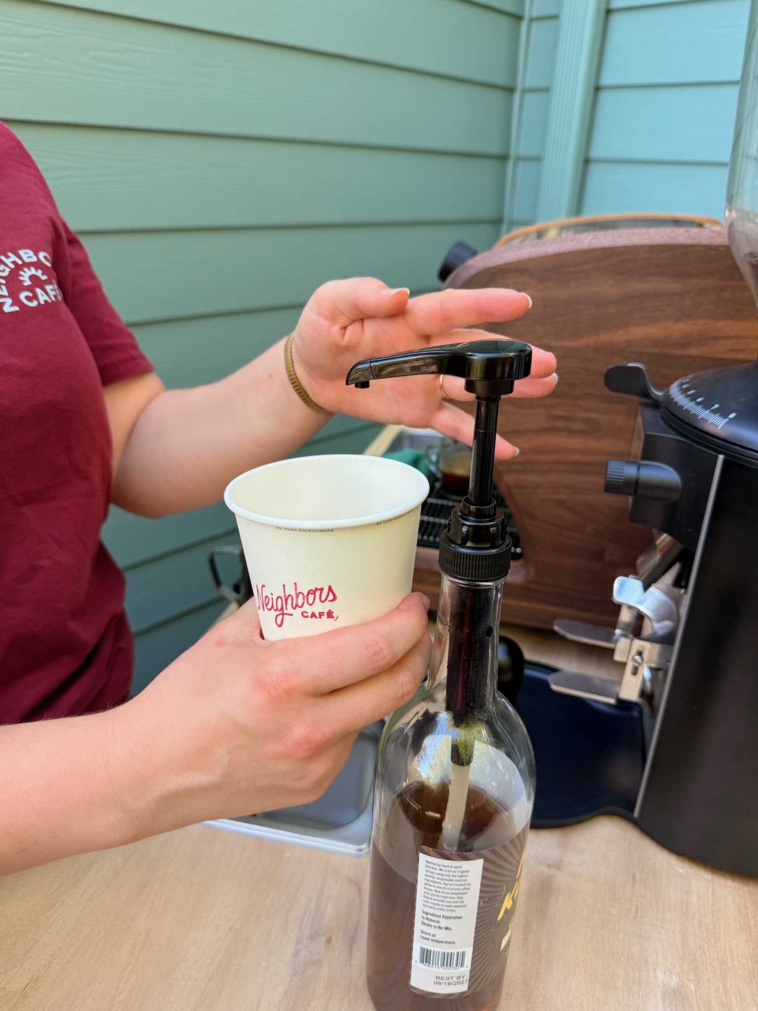 Person pouring chocolate syrup into a clear plastic bottle placed upside down on a table, with a Neighbors Café cup in hand, and a coffee machine in the background.