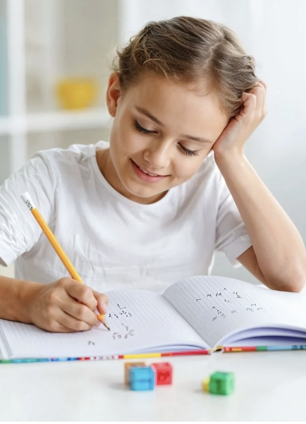 A young girl studying math in a notebook with colorful blocks on the table.