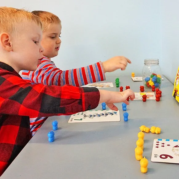 Two young boys playing a board game with colorful game pieces at a table, with a jar of multicolored game pieces in the background.