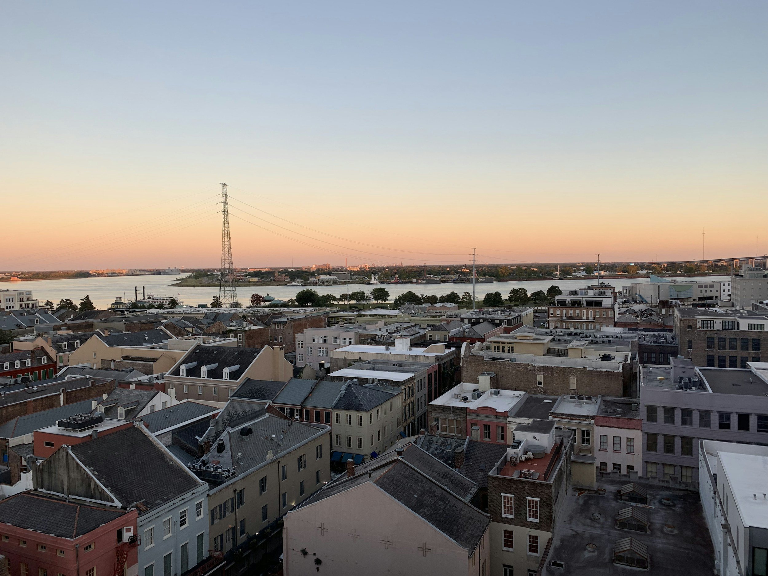 View of cityscape showing residential buildings with rooftops, a river with boats, and a distant horizon with industrial structures and communication towers during sunset. New Orleans cityscape highlighting residential neighborhoods and real estate