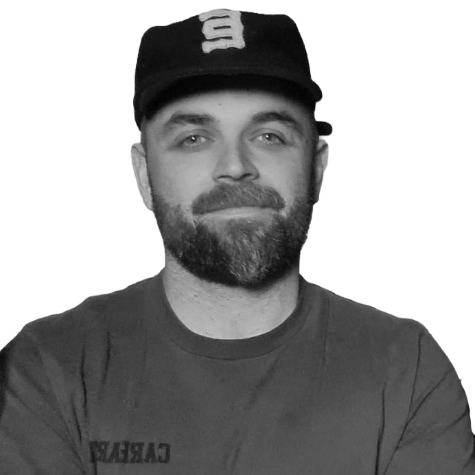 A man with a beard wearing a baseball cap and t-shirt, smiling slightly, with a plain background.