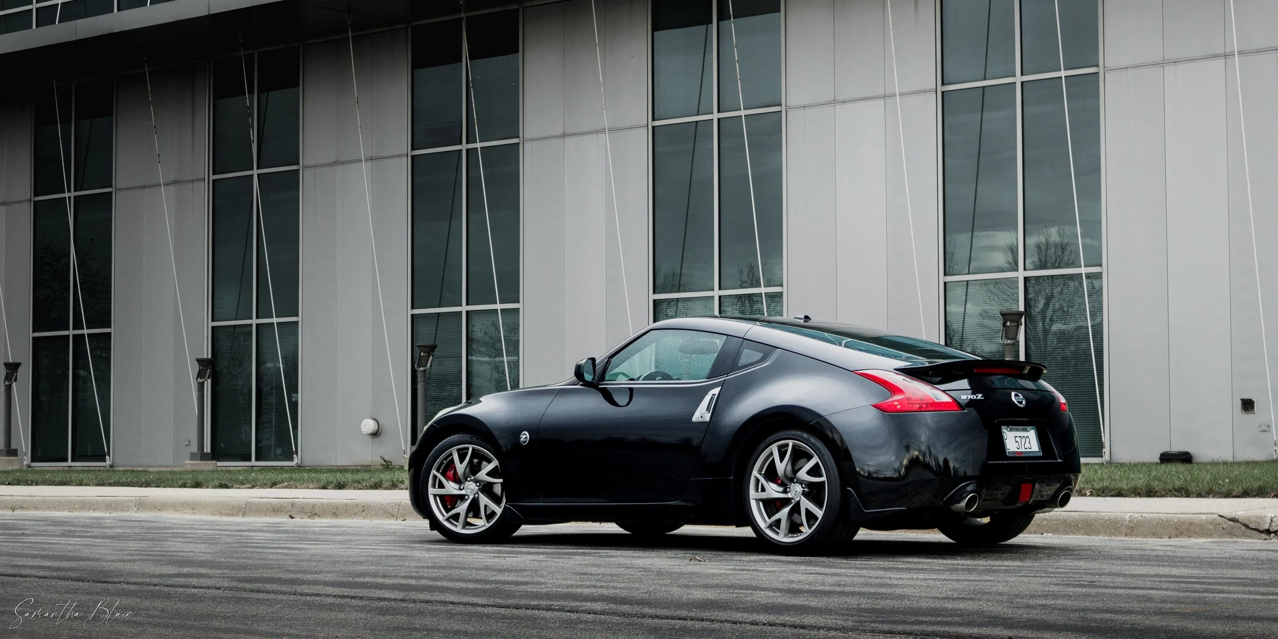 A black Nissan 370Z sports car parked on the street in front of a modern building with large glass windows.