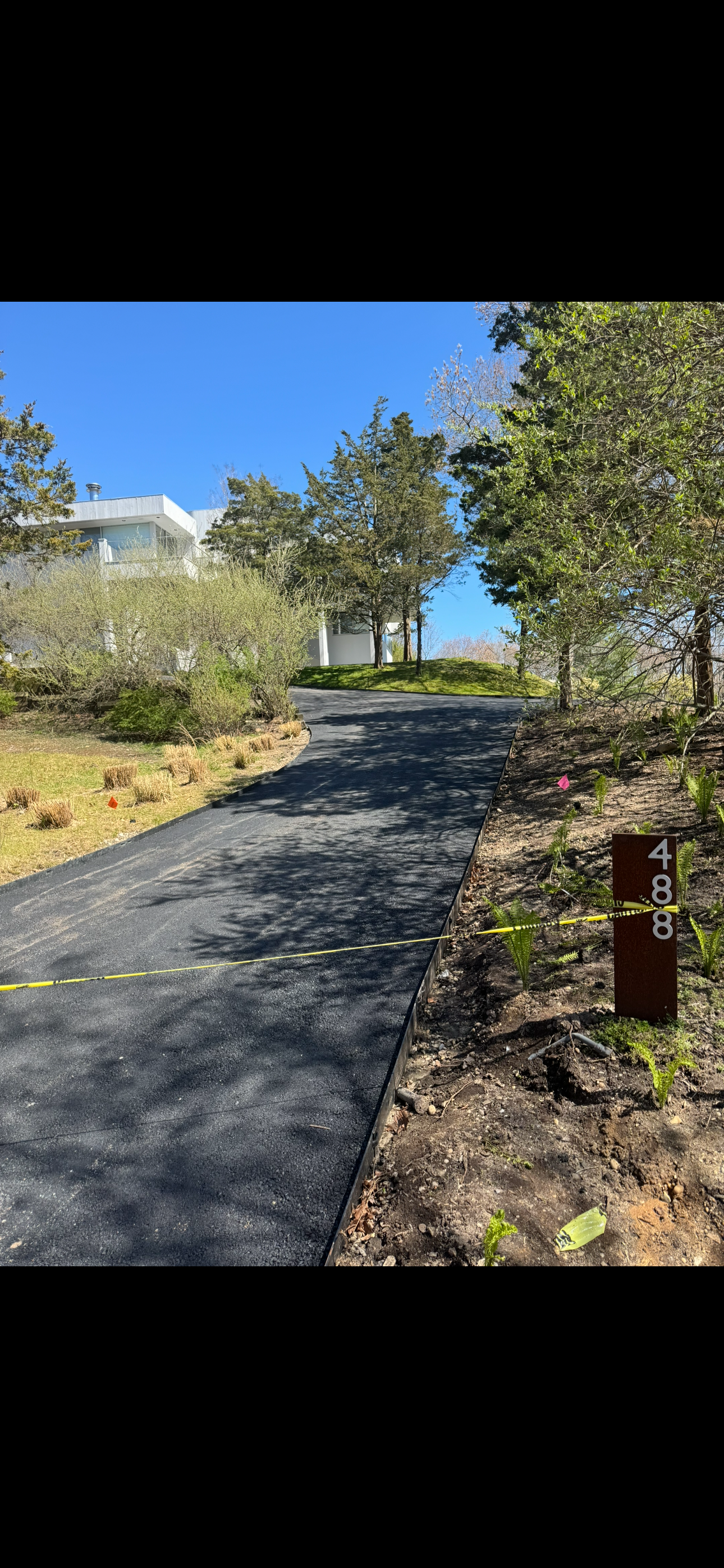 Freshly paved black asphalt driveway on a slight incline leading to a modern house with white walls and large windows. Surrounding greenery includes trees and small plants. A brown pole on the right has the number 488, and a yellow caution tape runs across the driveway.