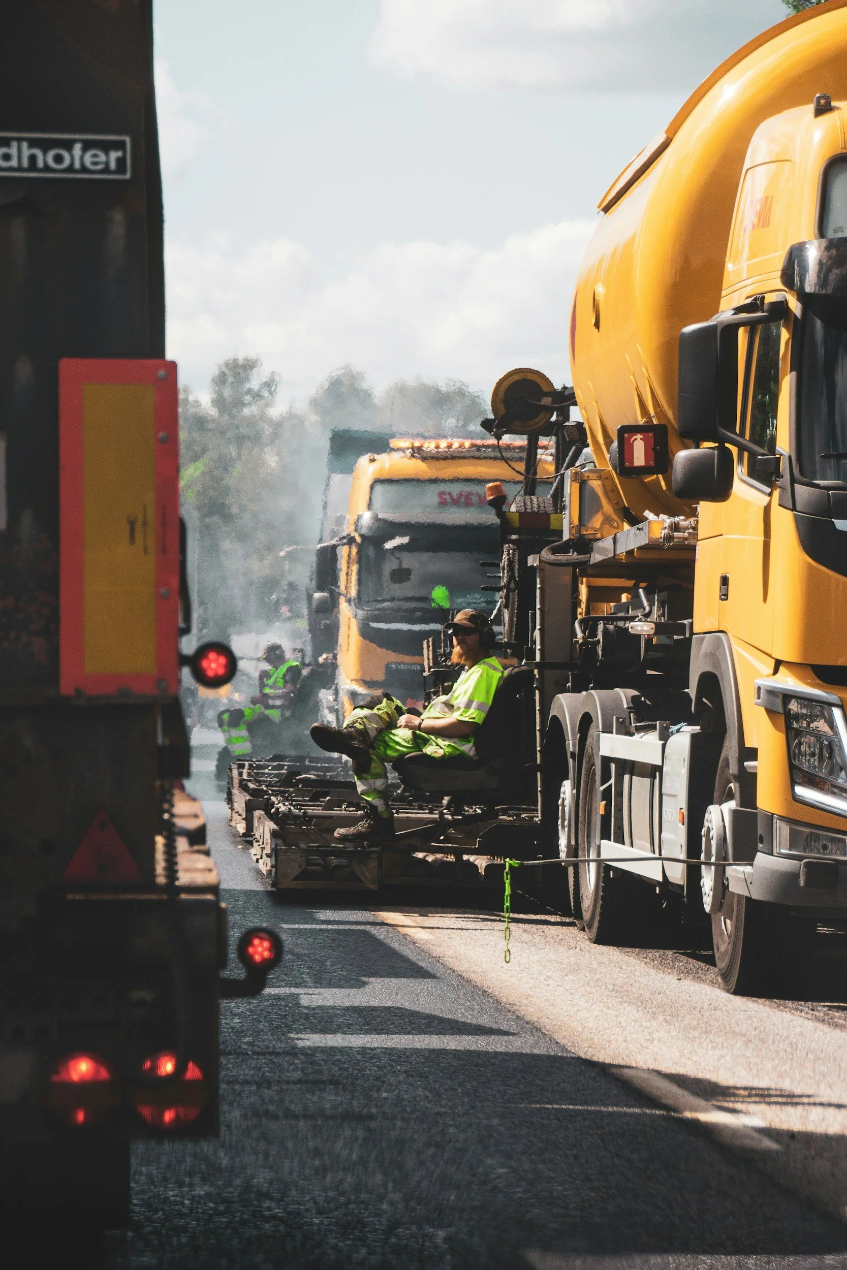 Construction workers in safety vests working on a road, with large yellow trucks and equipment around, in bright daylight.