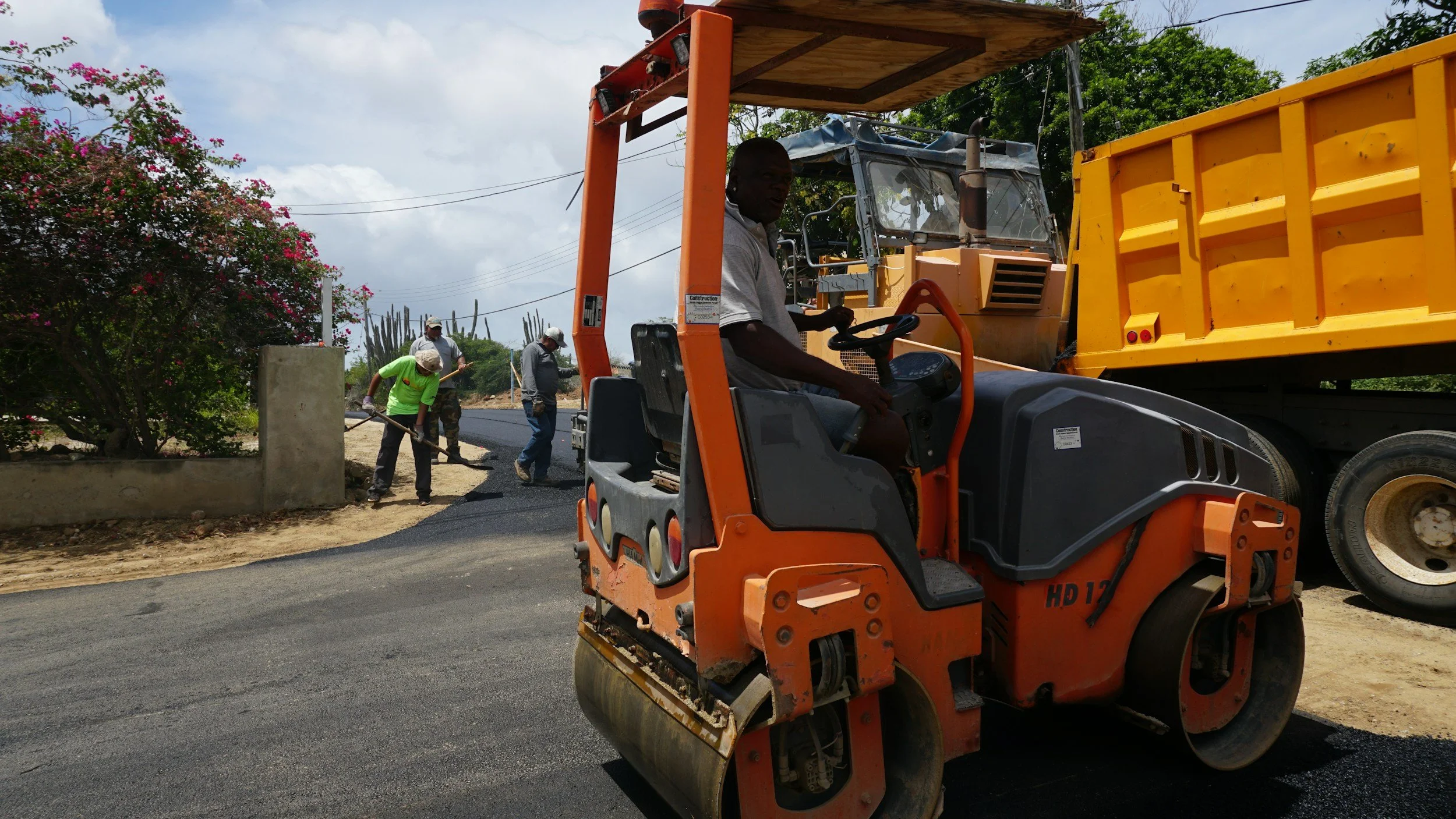 Workers are paving a road with heavy construction equipment, including a road roller and a dump truck, on a sunny day with trees and pink flowering bushes in the background.