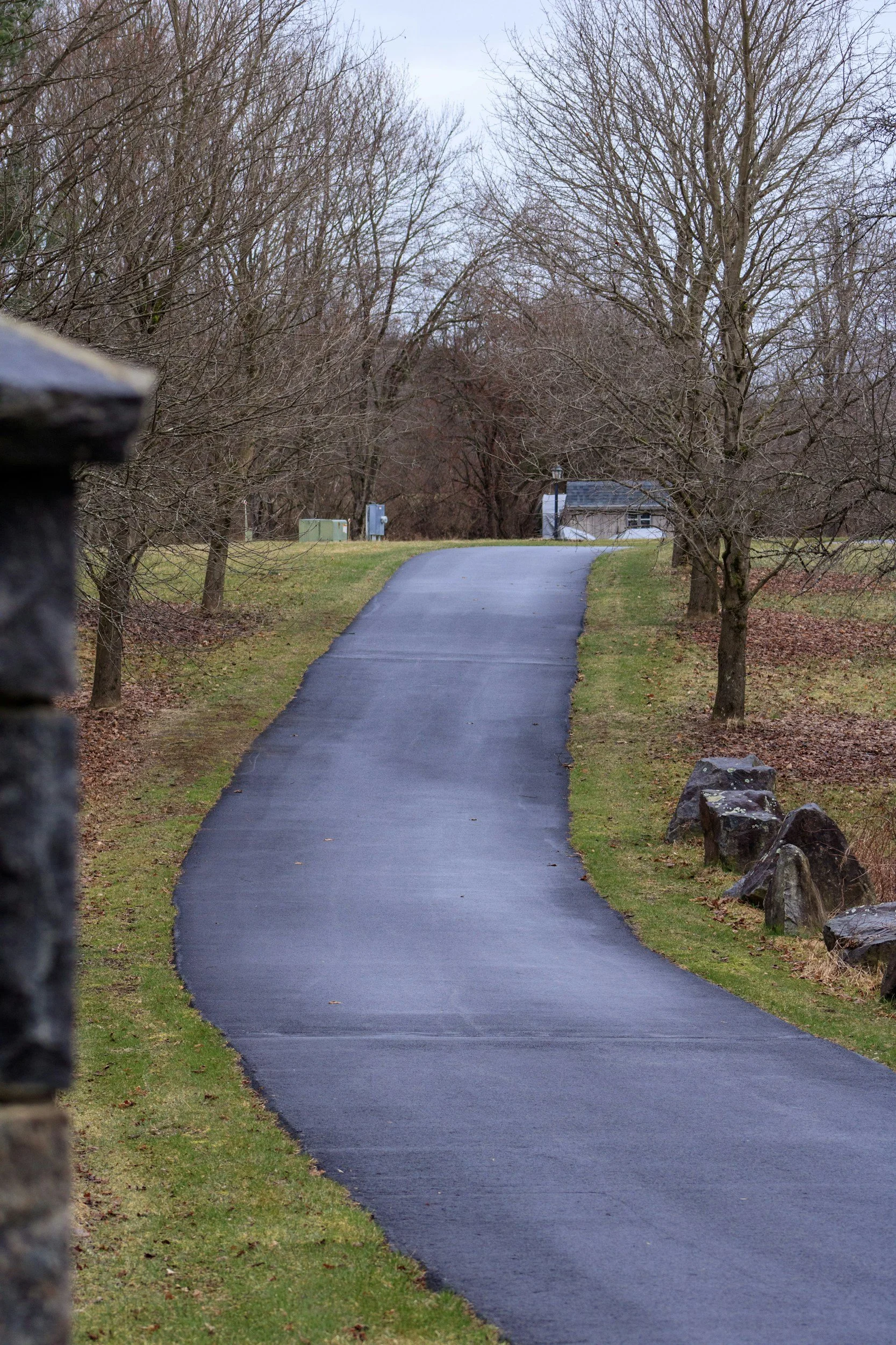 A paved winding path in a park during late fall or winter, bordered by grass, leafless trees, and large rocks on the right side; overcast sky.