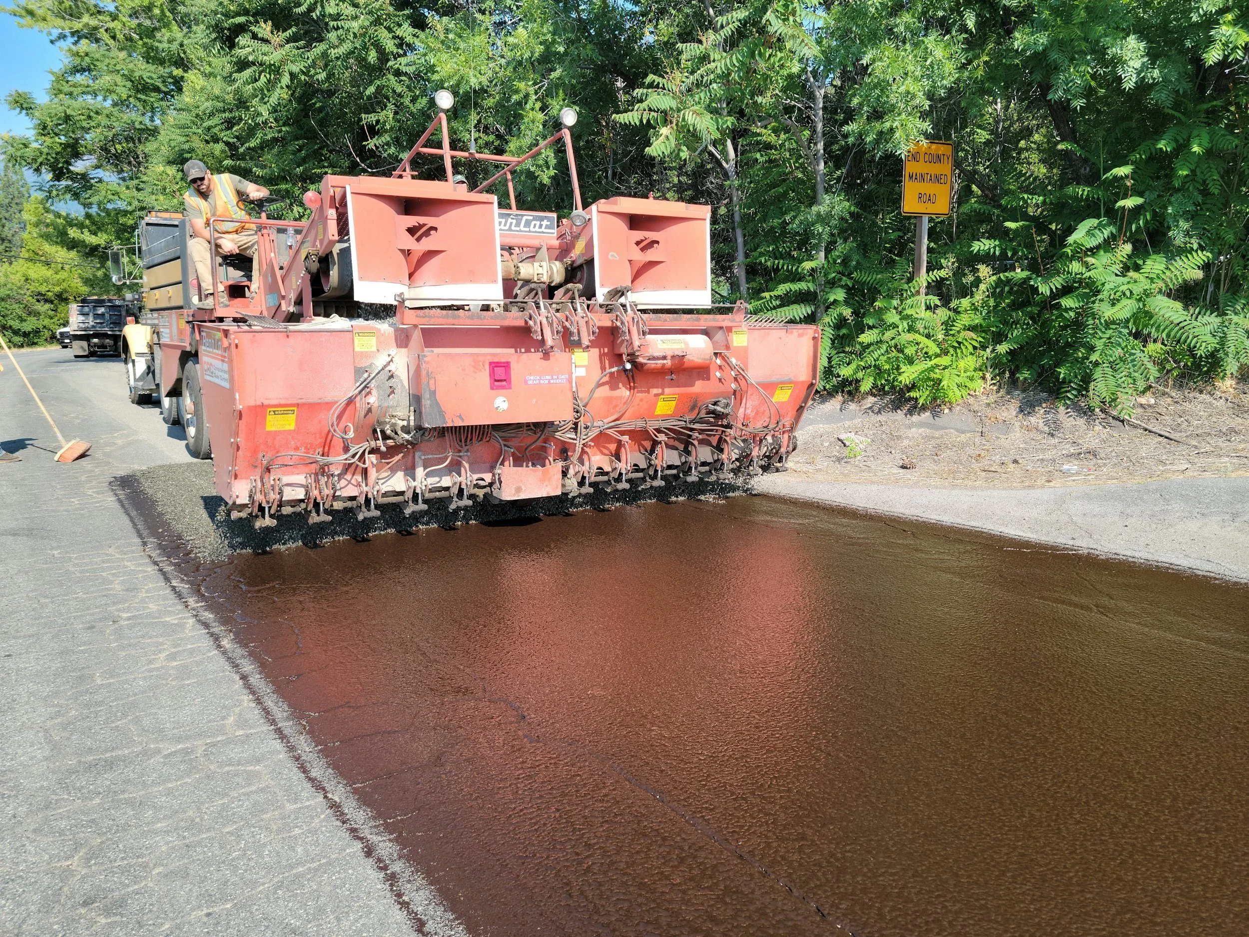 A construction worker operating a road paving machine on a sunny day, with trees and a yellow road sign that reads 'End County Maintained Road' in the background.