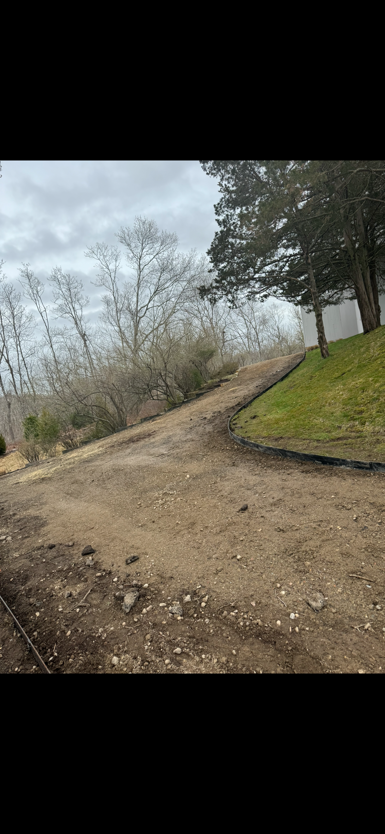 A dirt pathway curves around a grassy area with trees in the background, under a cloudy sky.