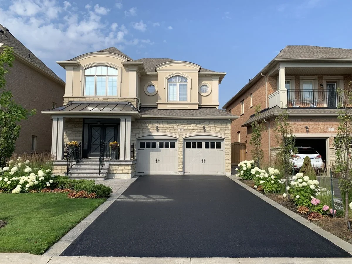 A two-story house with stone and beige siding, two garage doors, steps leading to a front porch, and floral landscaping on both sides of the driveway.