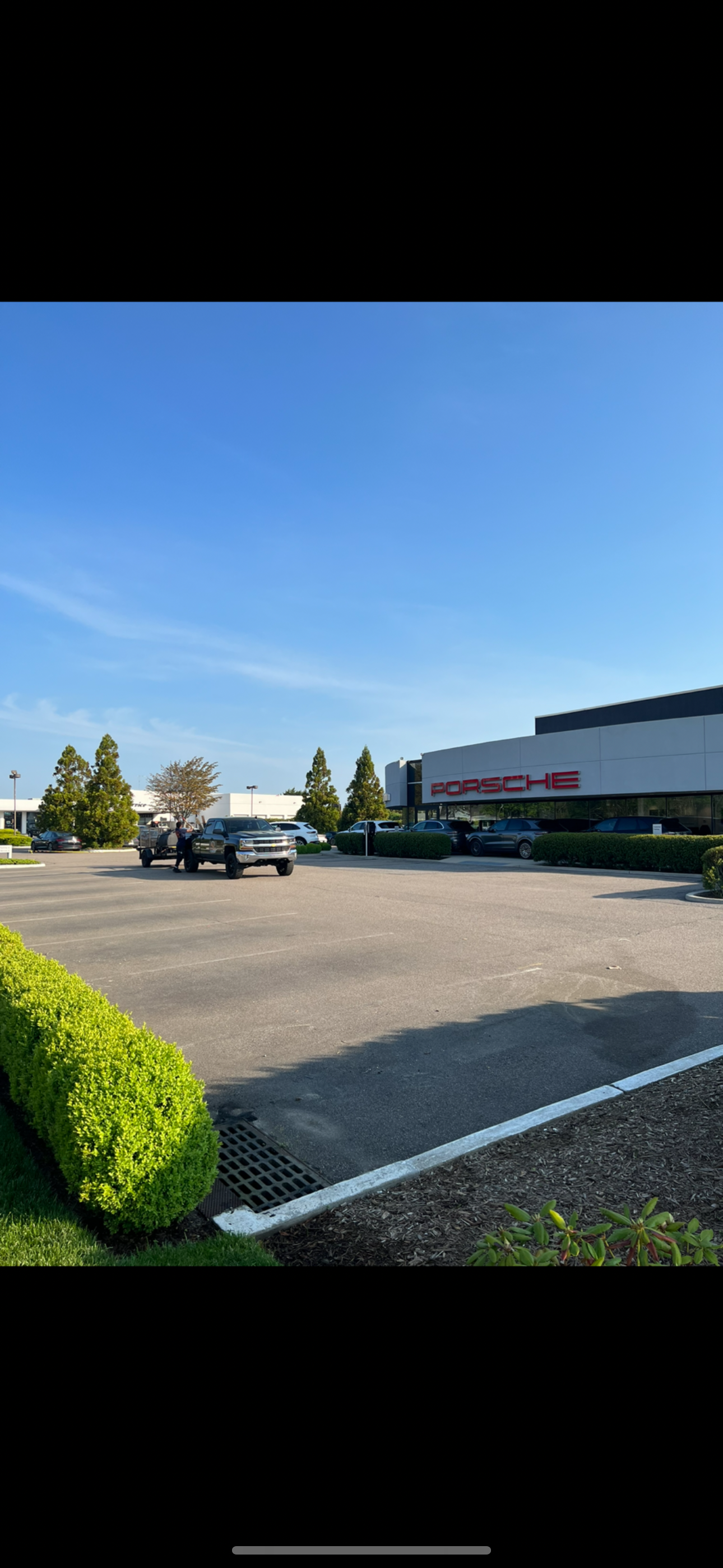 View of a Porsche dealership with cars parked in the lot, people near a truck, trees, and a building with the Porsche logo under a clear blue sky.