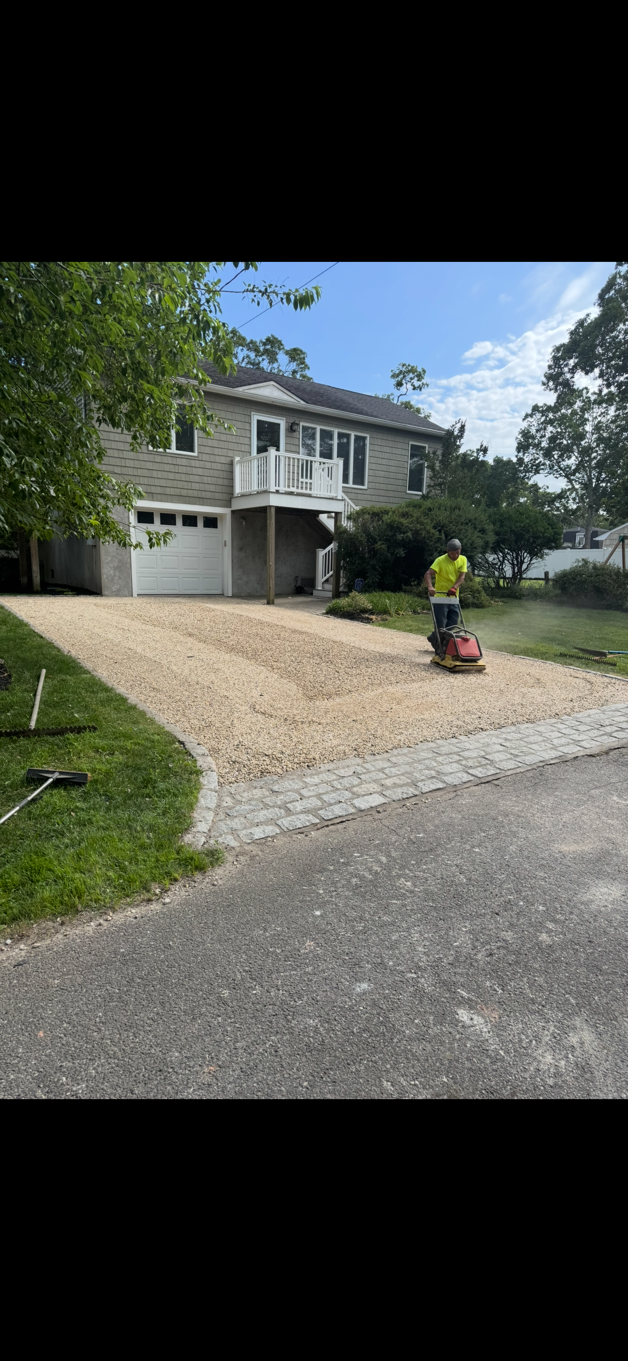 A person wearing a yellow shirt is using a compactor on a gravel driveway in front of a gray house with a garage and a balcony.