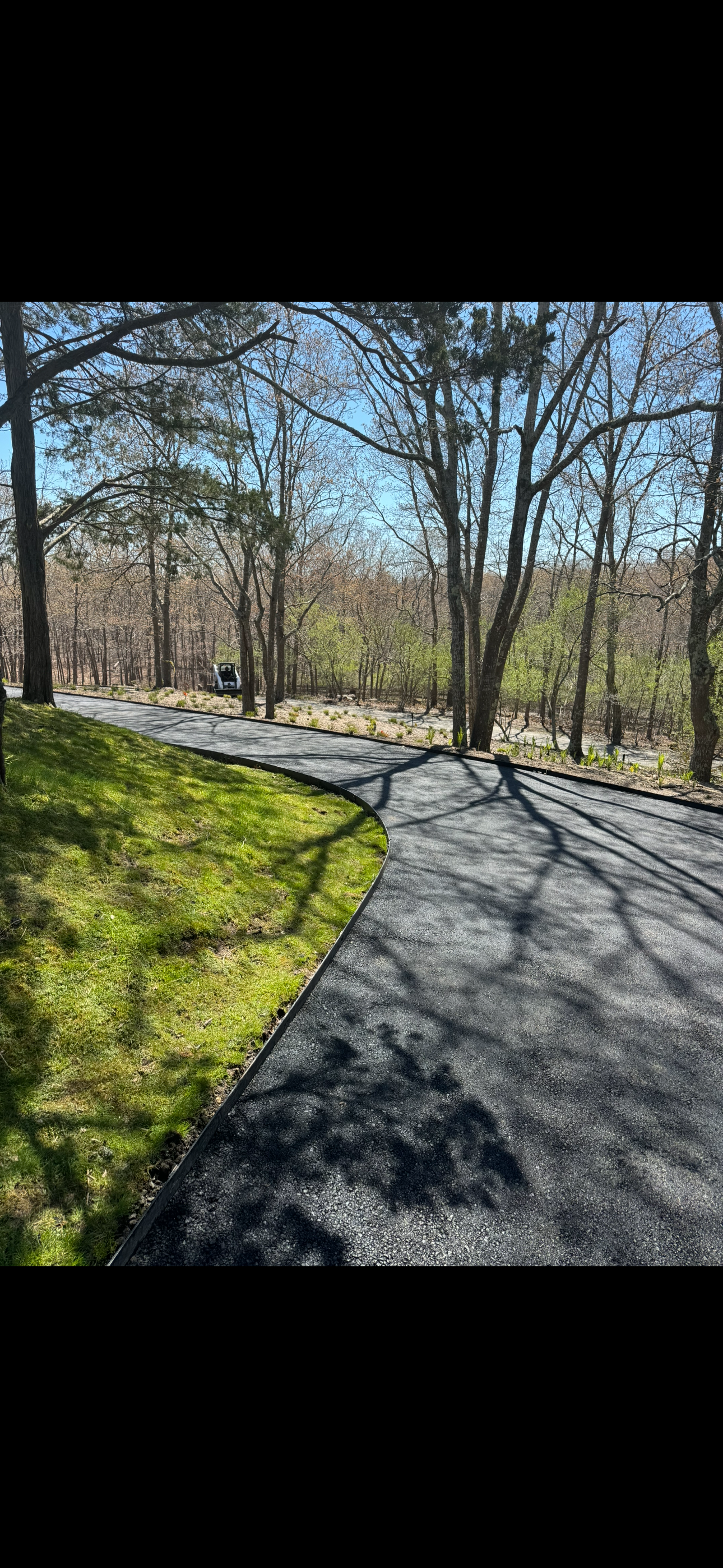 Curved newly paved asphalt driveway with shadows of tree branches, green grass on the side, in a wooded area with leafless trees and a clear blue sky.