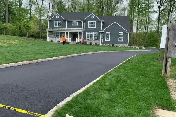 A newly paved asphalt driveway leading to a large gray house with white trim, surrounded by a lush green lawn and trees.