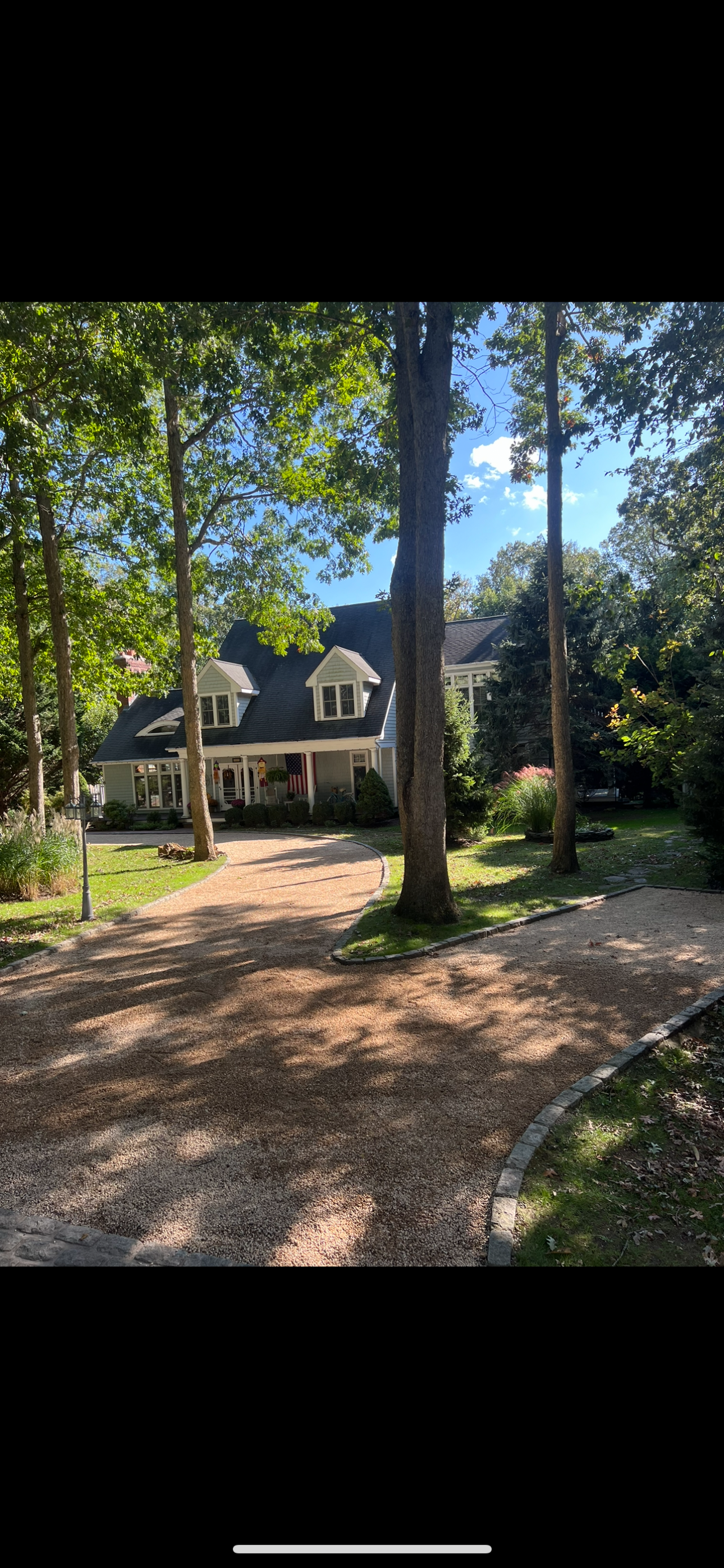 A house with a black roof and white walls, surrounded by tall trees and greenery, with a curved gravel pathway leading to the entrance, sunny weather, and a blue sky with some clouds.