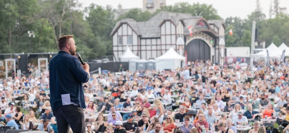 A man speaking into a microphone at an outdoor comedy performance with a large crowd of people in Winnipeg.