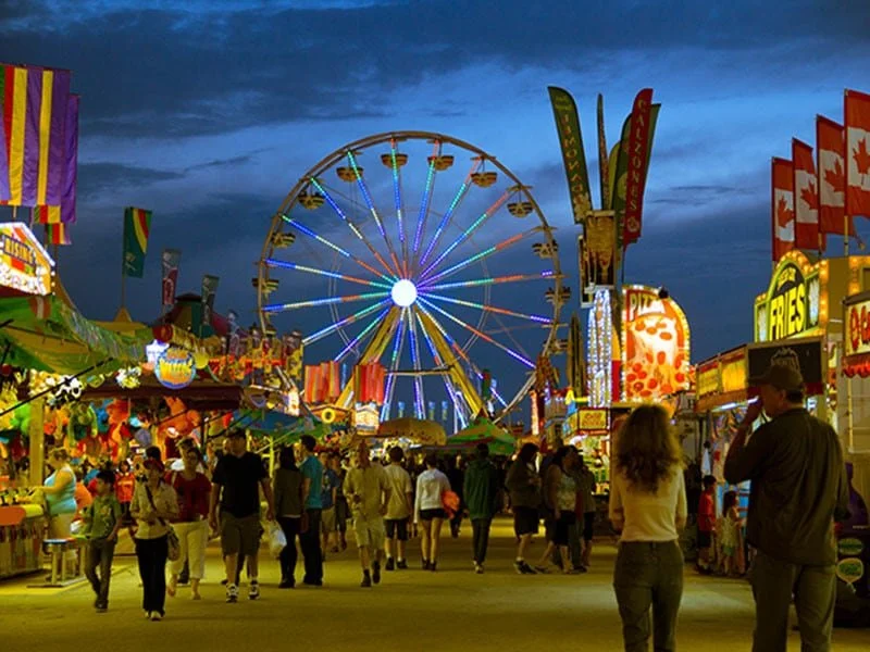 People walking at a colorful carnival with a large Ferris wheel and various game and food booths, under a cloudy evening sky in Winnipeg.