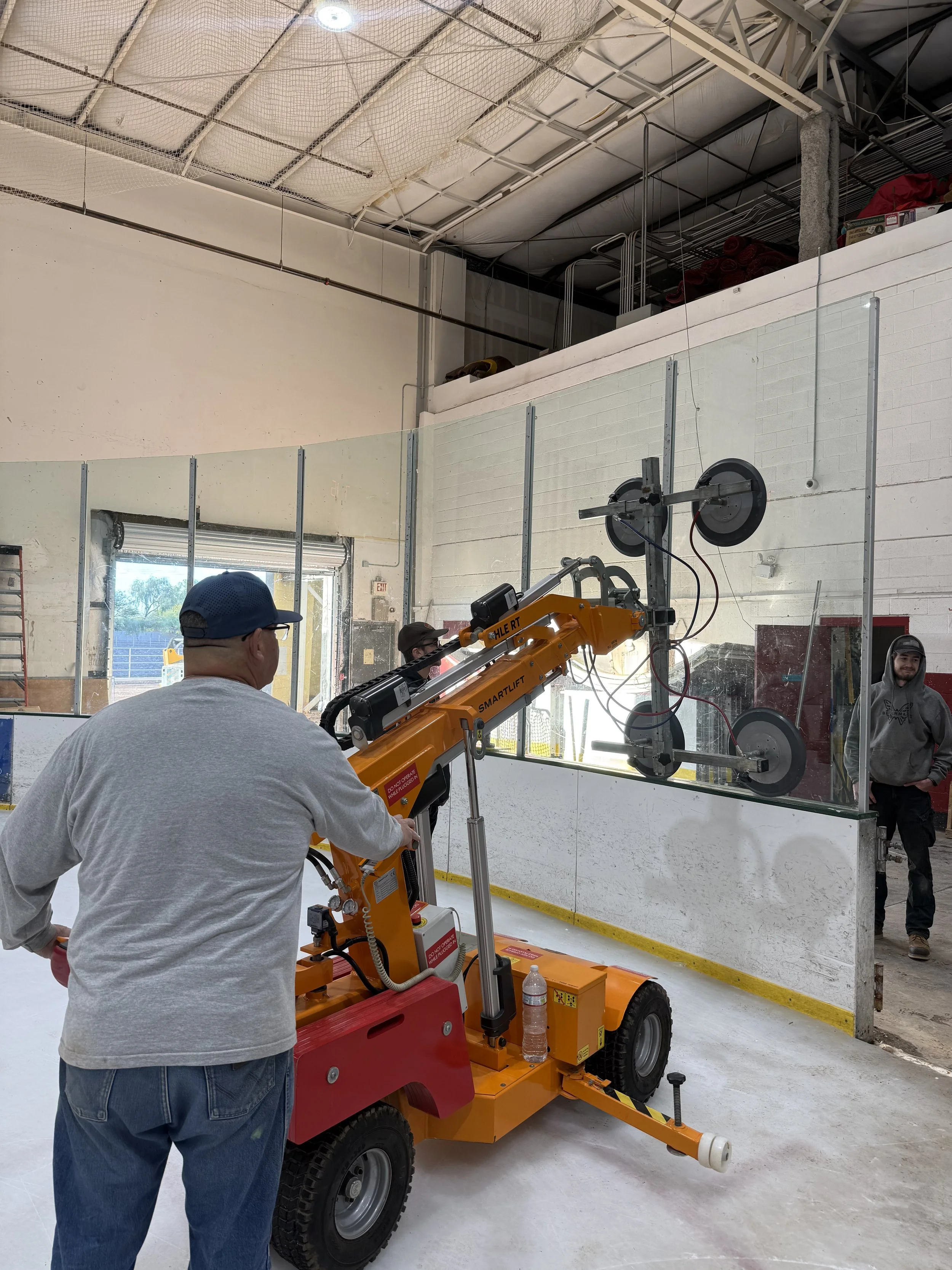A person operating a glass handling robot in an ice skating rink, with a man in a hoodie observing nearby.
