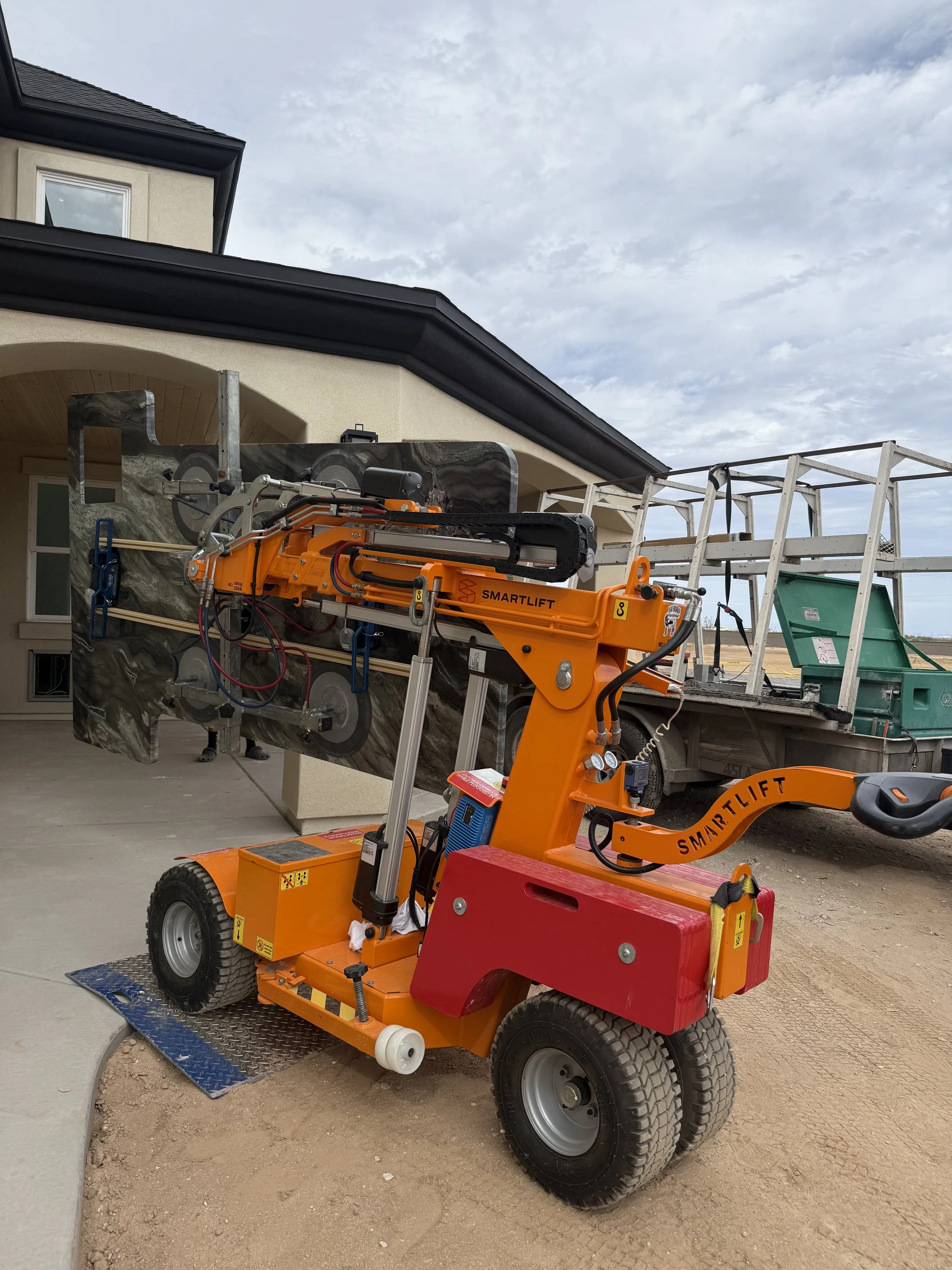 A construction site with a small orange and red robotic lift in the foreground, a house with beige walls and black roof in the background, and a cloudy sky overhead.