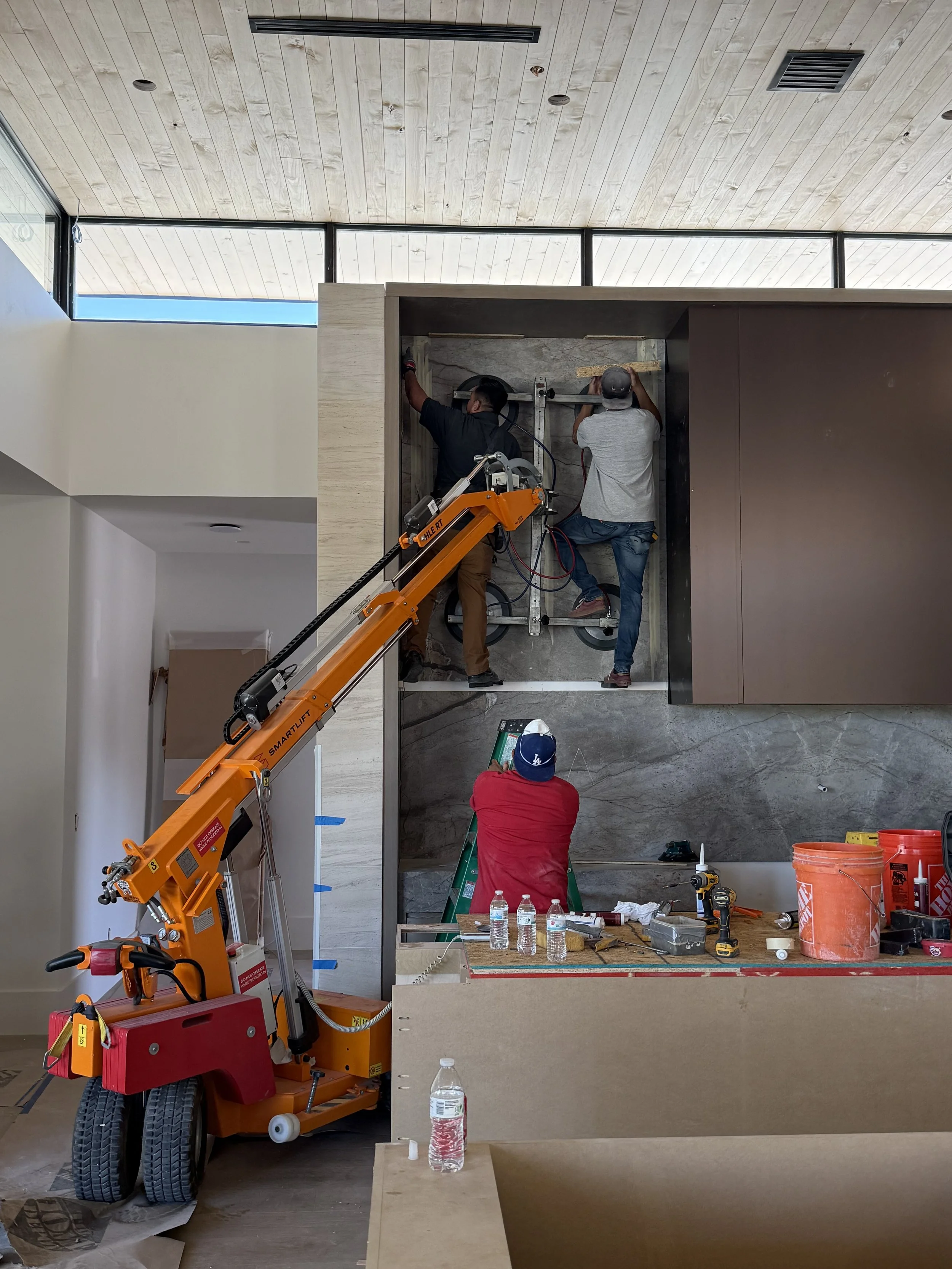 Three workers installing or repairing a large wall panel in an indoor space. One worker is on a ladder, and two are on a scissor lift. Various tools and water bottles are on a table nearby.